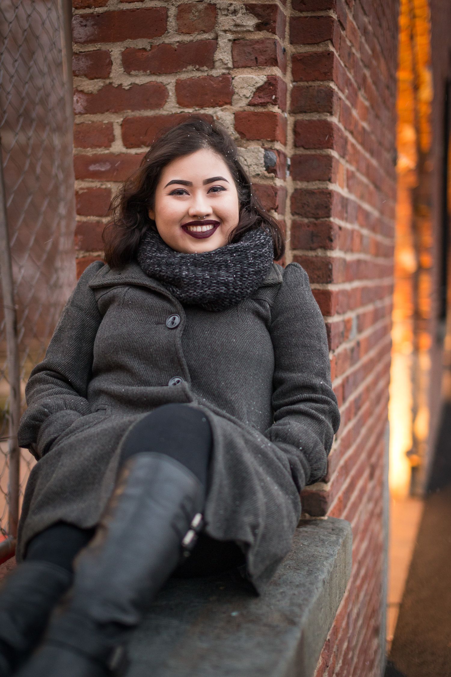 Young Asian woman smiling in gray coat and scarf seated by brick wall.