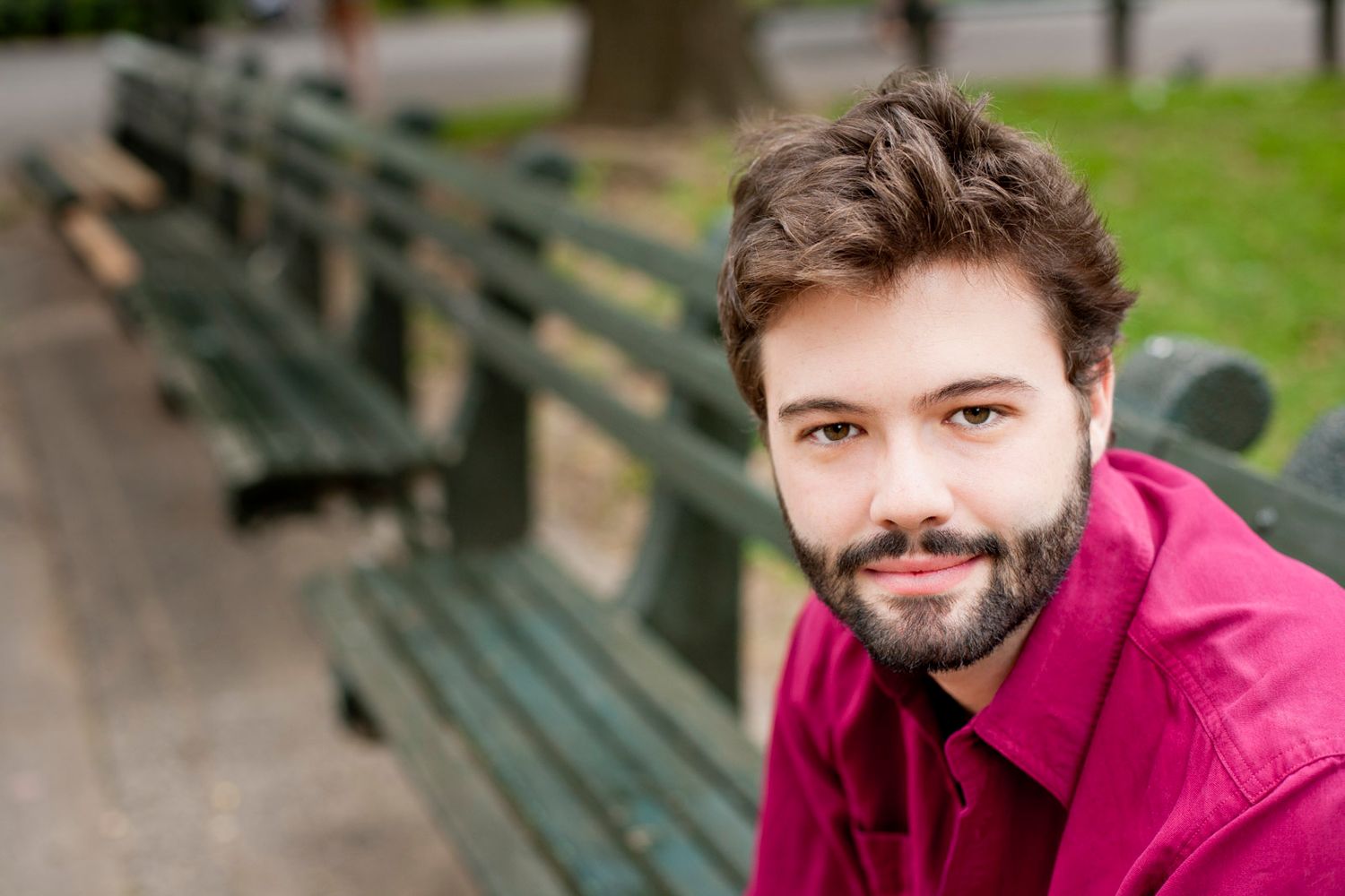 Young bearded man in pink shirt smiling on park bench outdoors.