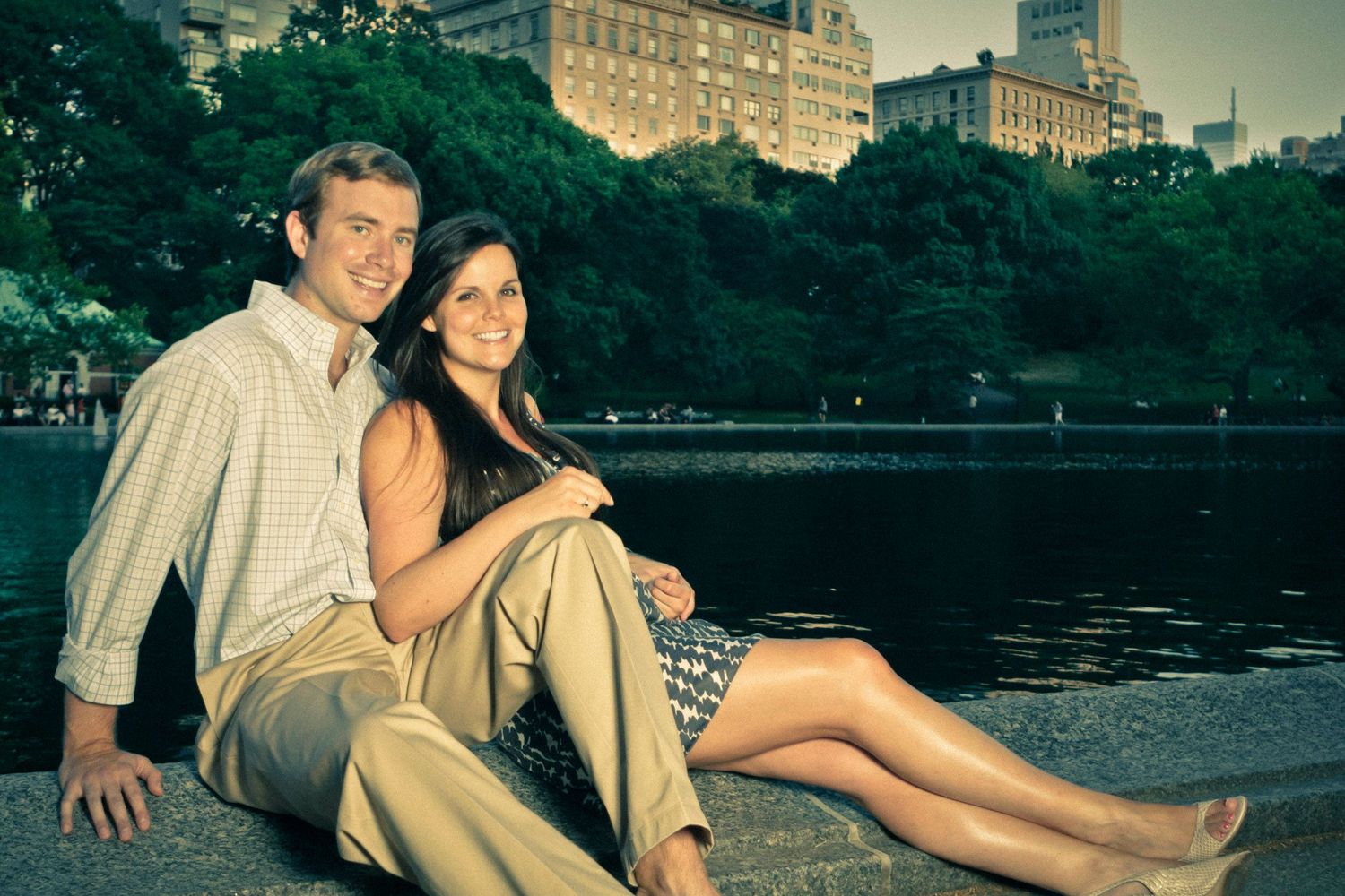 Smiling couple sitting closely together by a park pond at dusk with city buildings behind.
