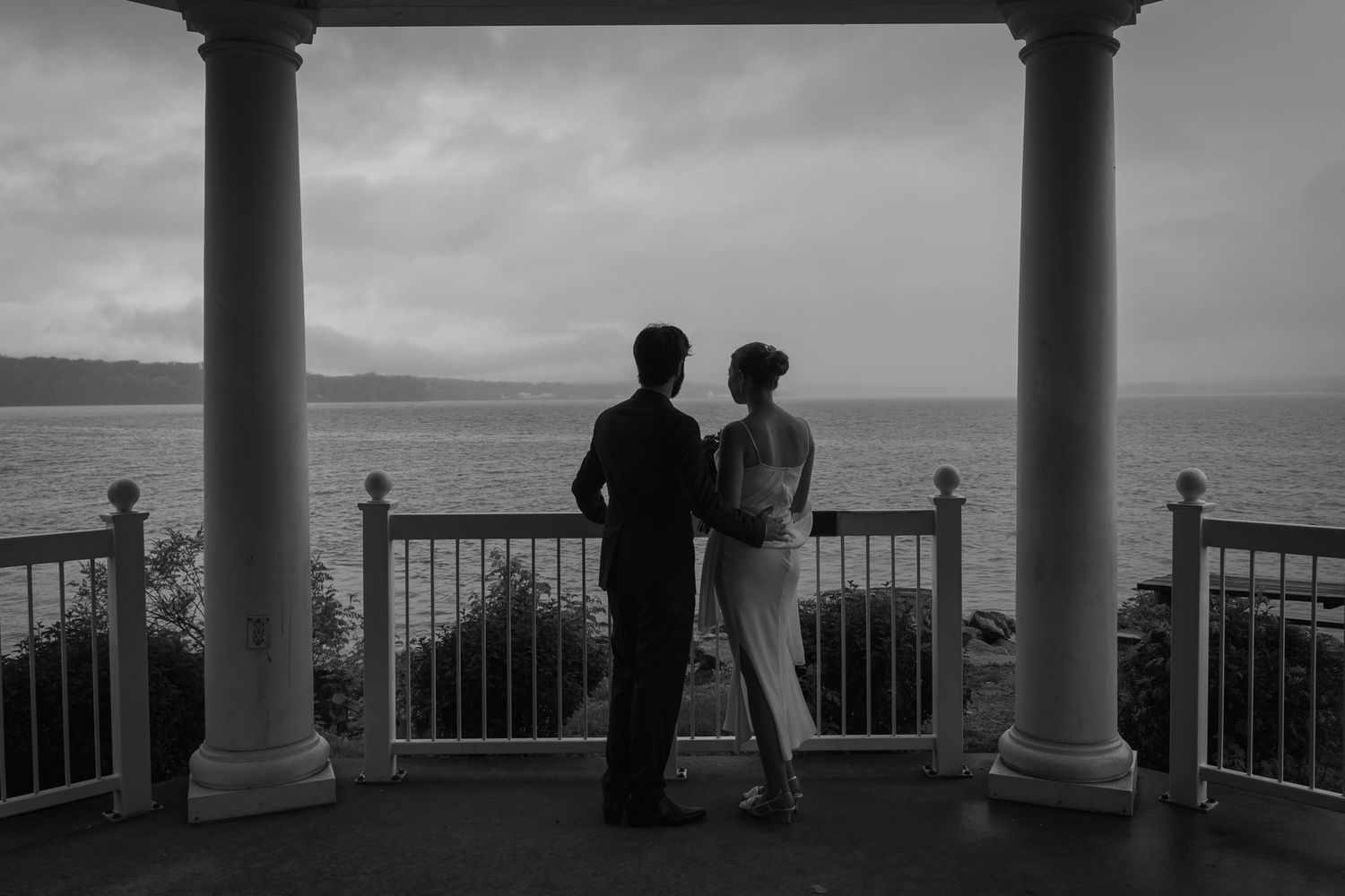 Black and white photo of a couple standing on an elegant columned veranda overlooking a misty ocean at dusk.