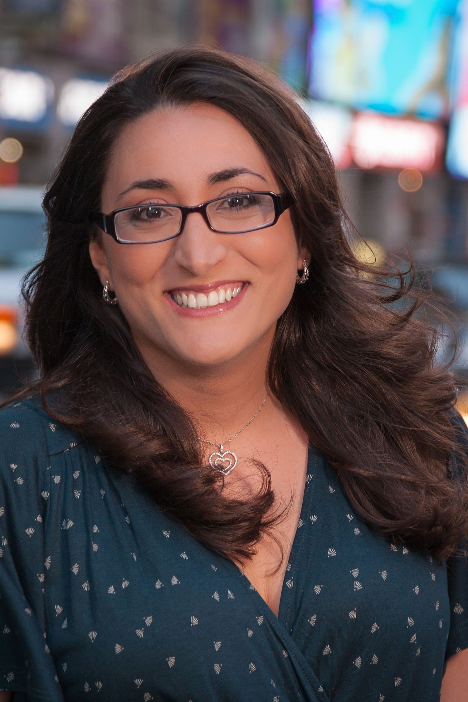 Smiling brunette woman with glasses and wavy hair wearing a teal blouse and heart necklace outdoors.
