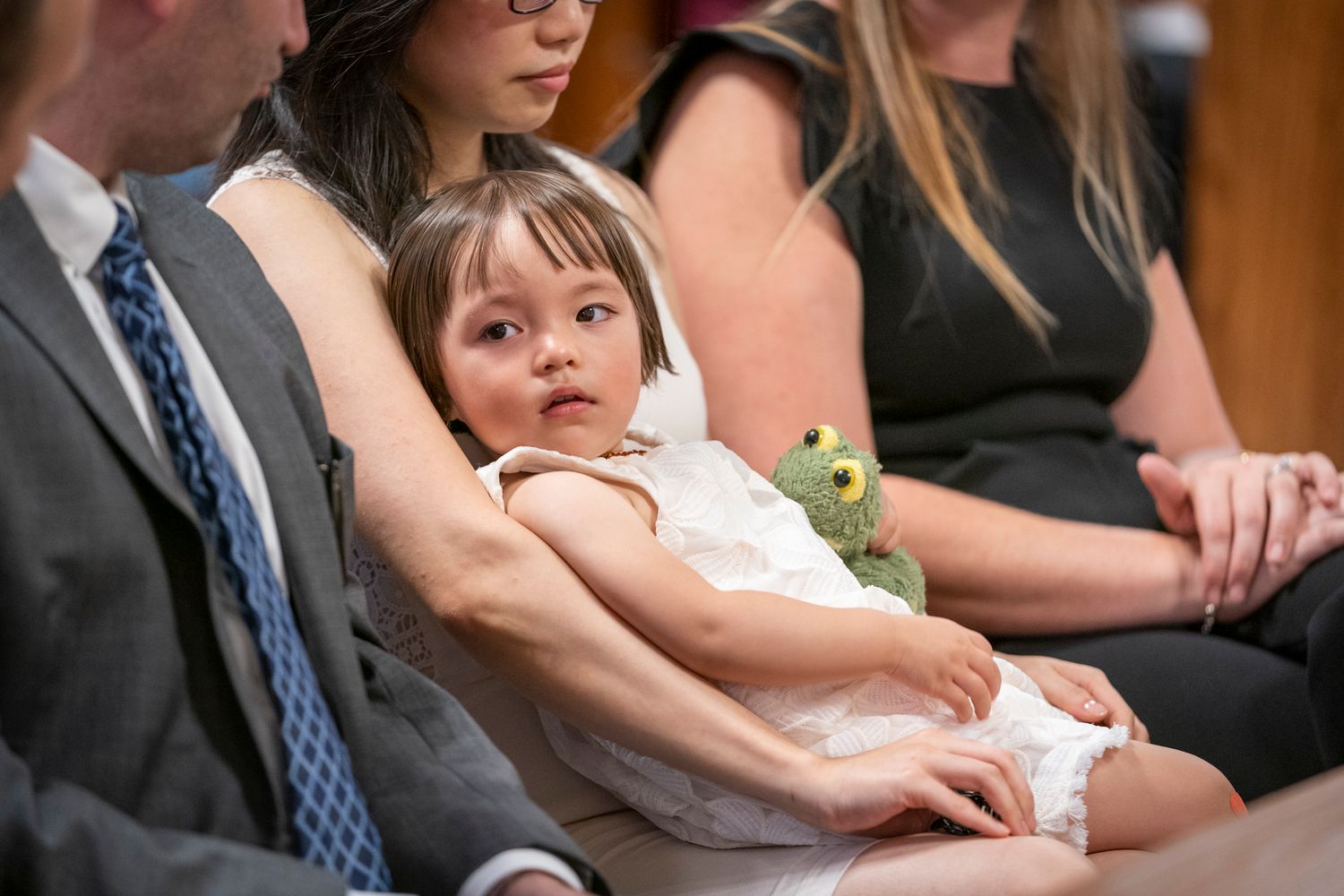 Young toddler girl in white dress holding green frog toy sitting on mothers lap during church ceremony.
