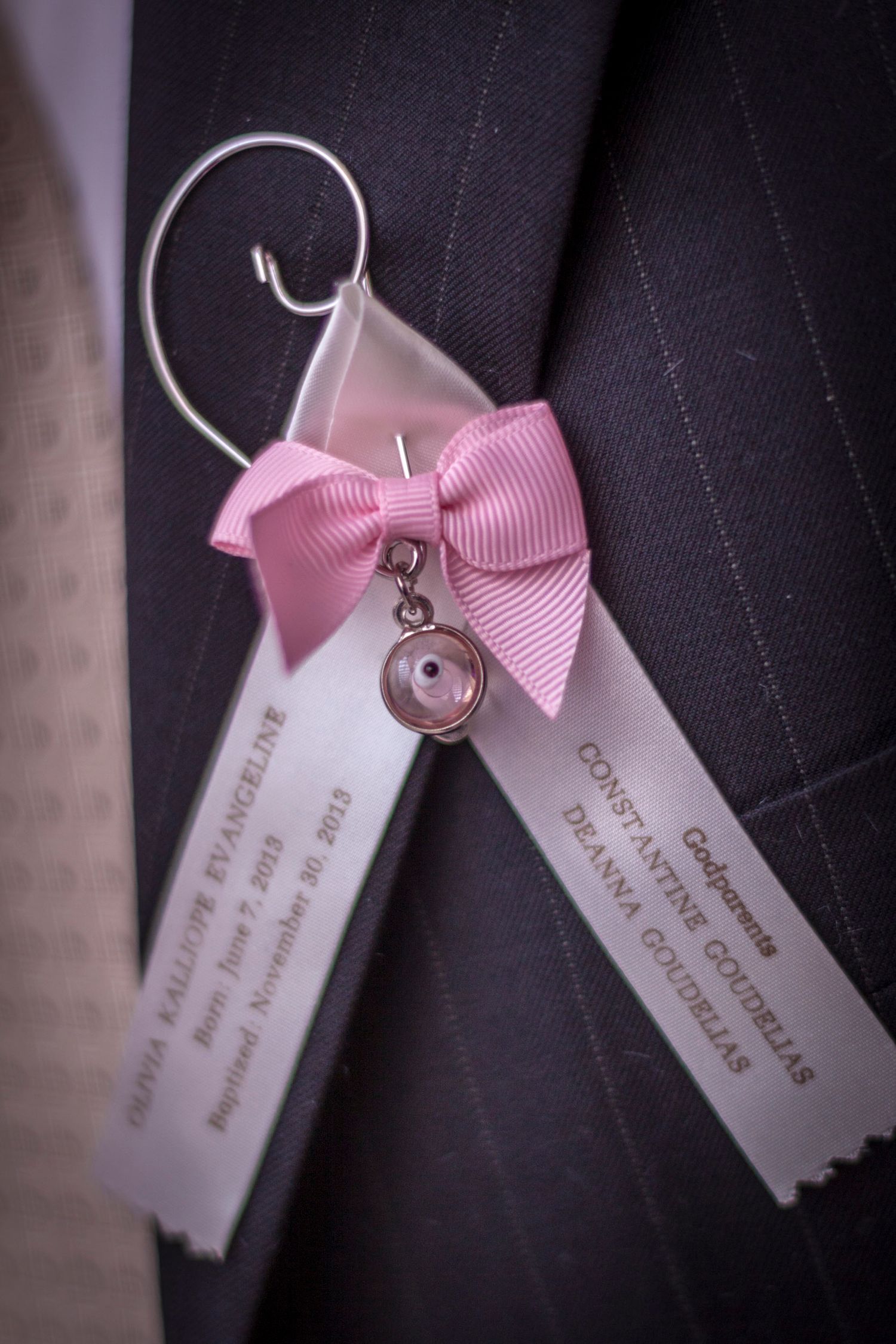 Pink bow boutonniere with ribbon tags and charm on pinstripe suit jacket for baptism ceremony.