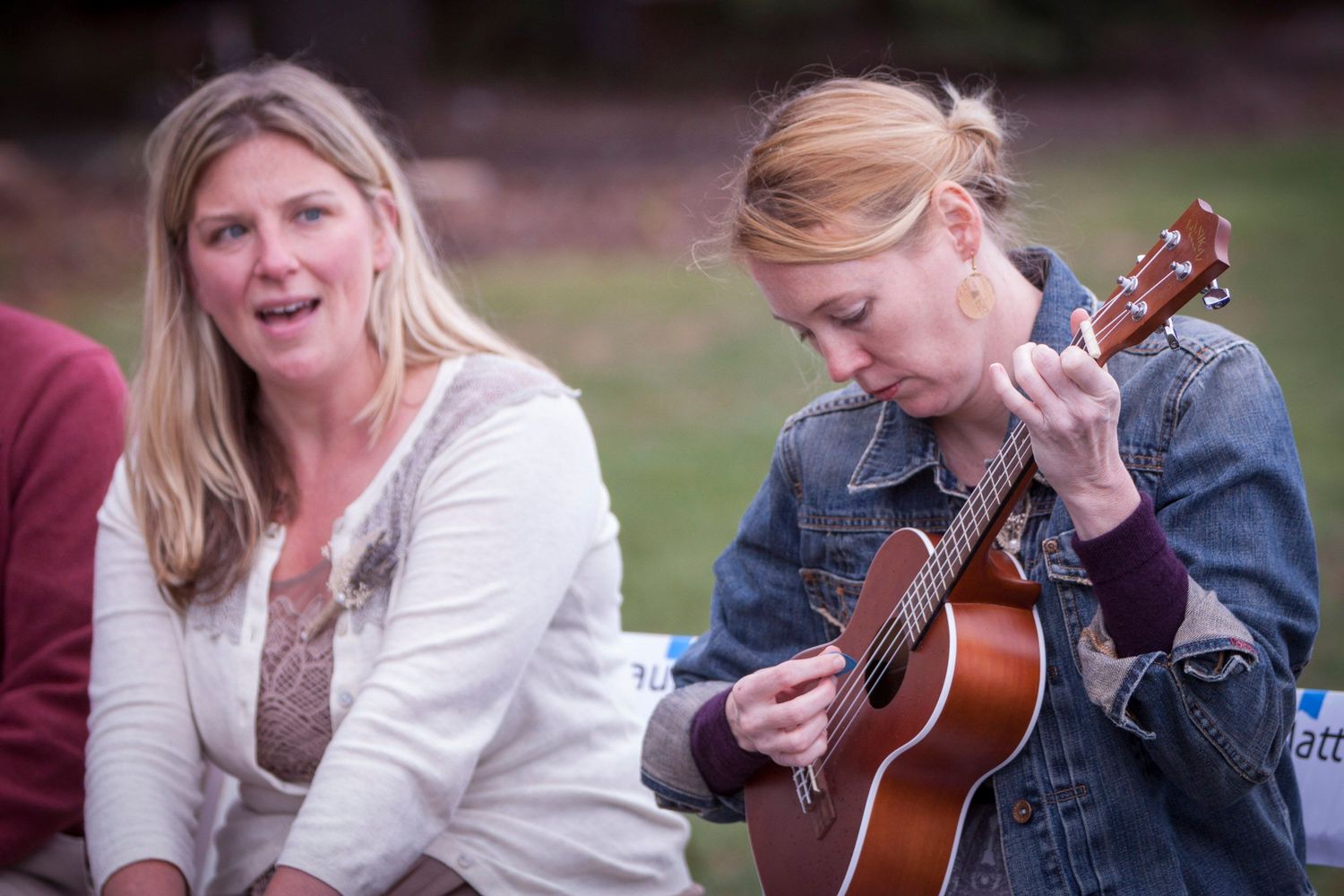 Two women outdoors, one playing acoustic guitar in denim jacket, other singing in white cardigan.