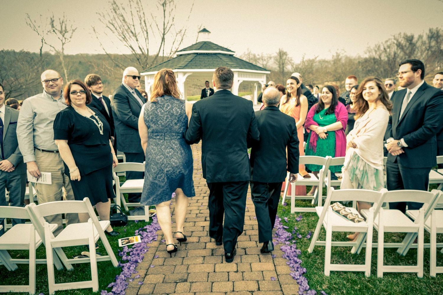 Outdoor wedding ceremony with guests standing along a brick aisle leading to a gazebo, surrounded by lush greenery.