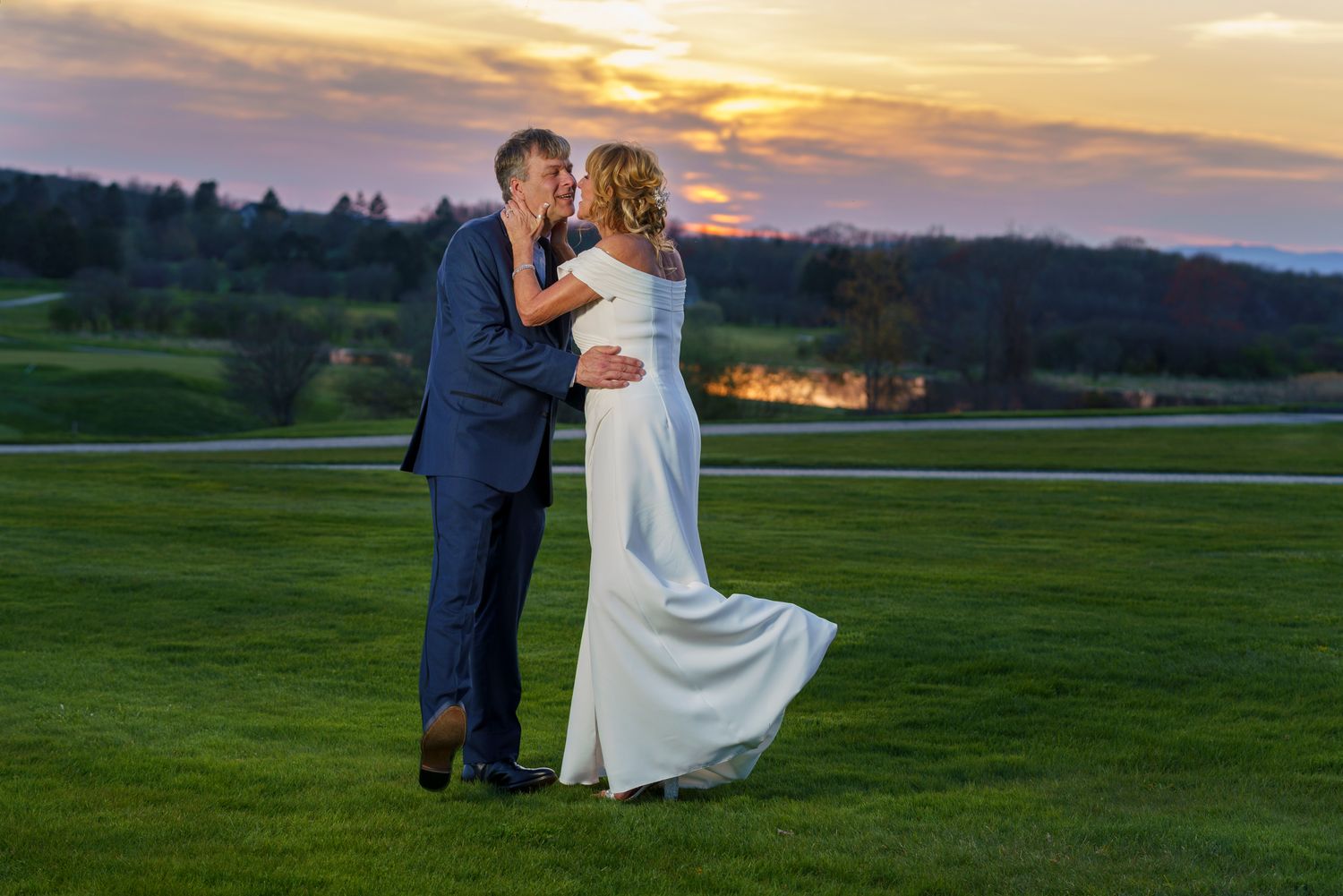 Bride and groom share a romantic kiss on a green lawn at sunset, with a glowing orange sky in the background.