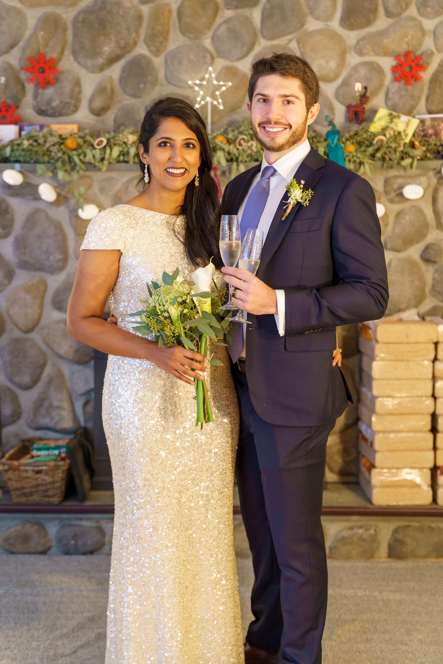 Bride in sequin gown holding bouquet and groom in navy suit toasting with champagne before stone fireplace.