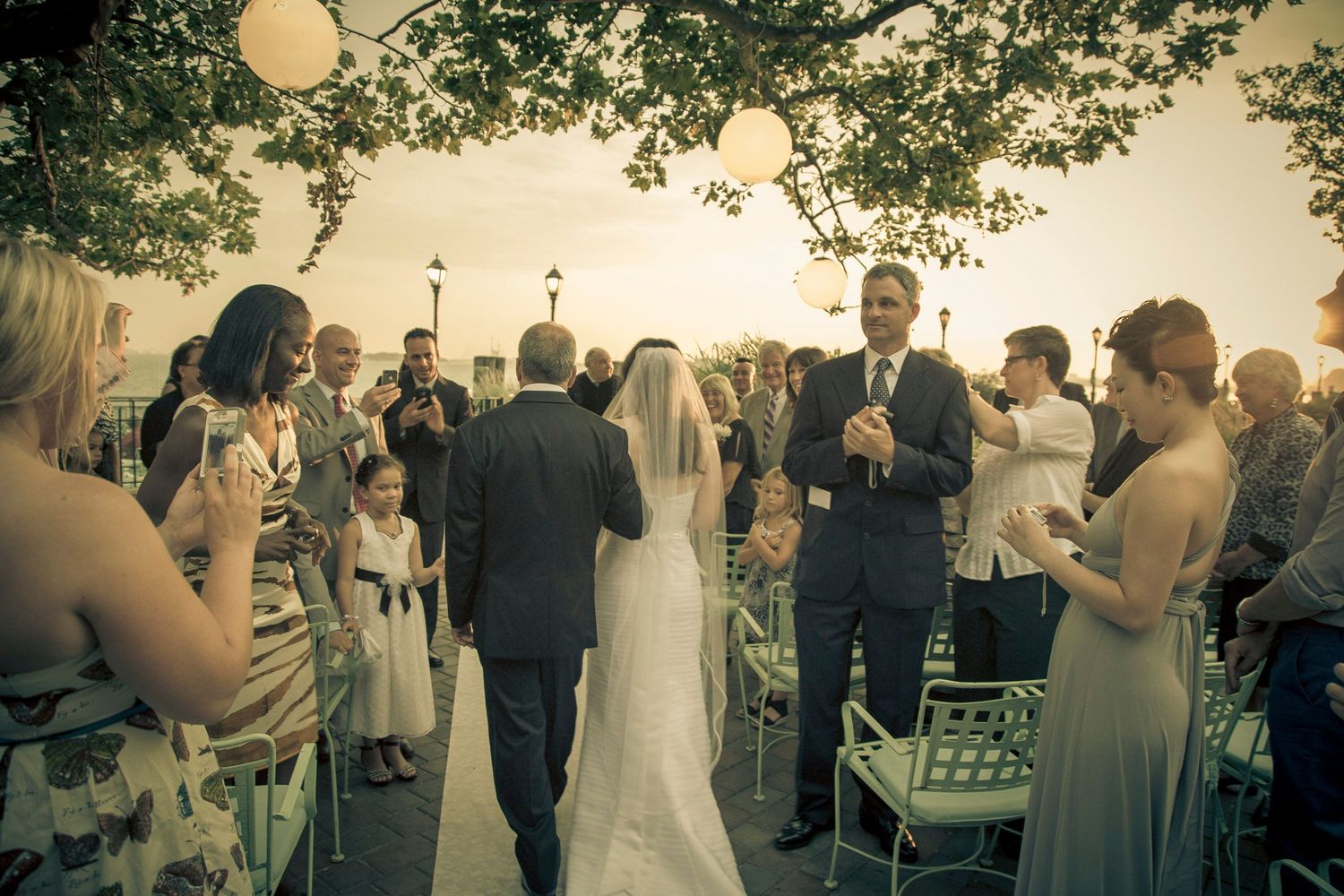 Bride walks down the aisle at an outdoor wedding ceremony with guests applauding.