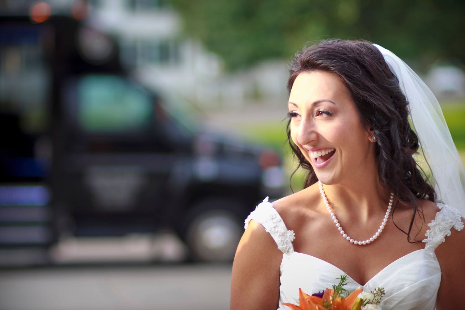 A happy bride in a white gown and veil holding an orange bouquet outdoors.