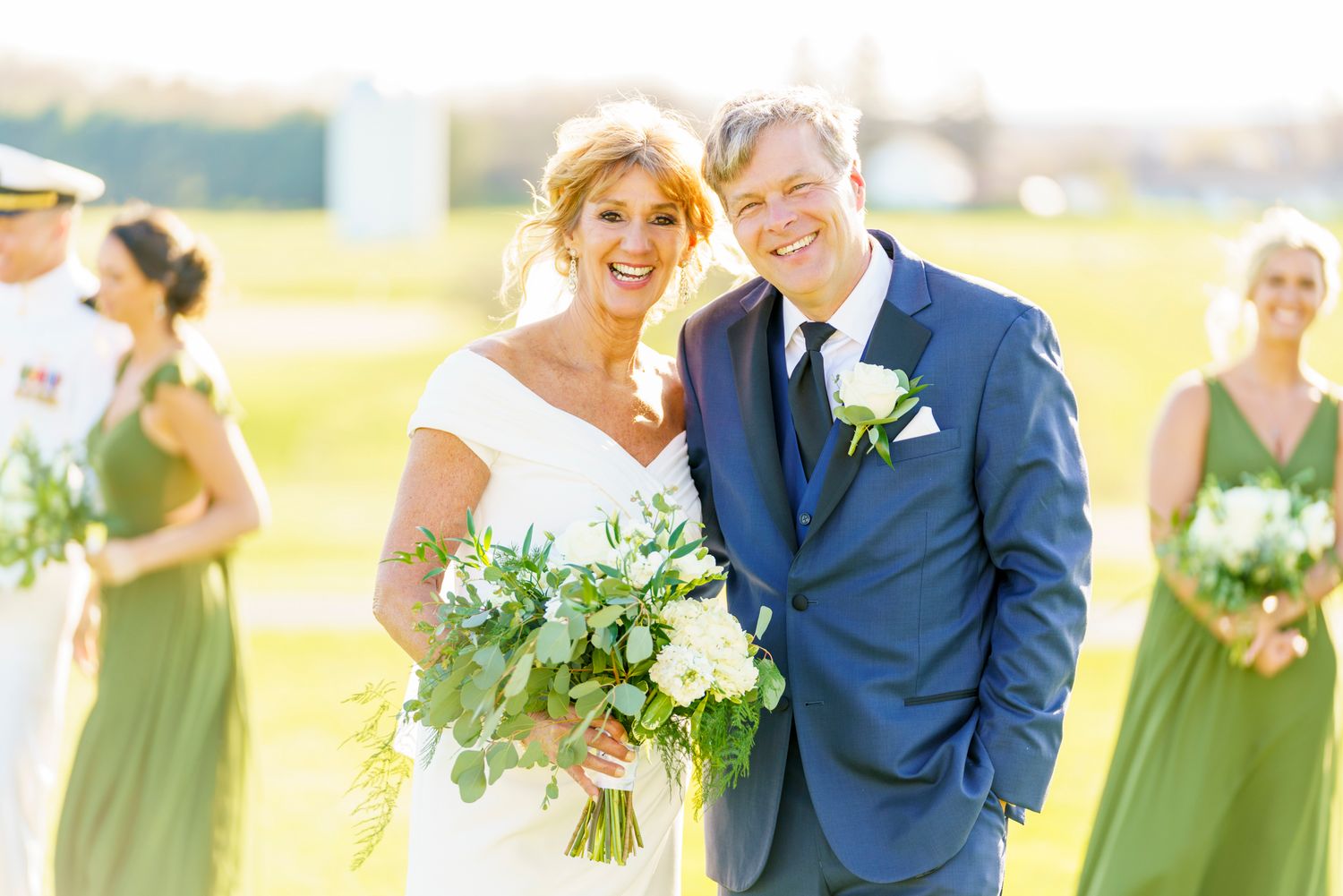 Smiling bride and groom with bridal party in sage green dresses outdoors.
