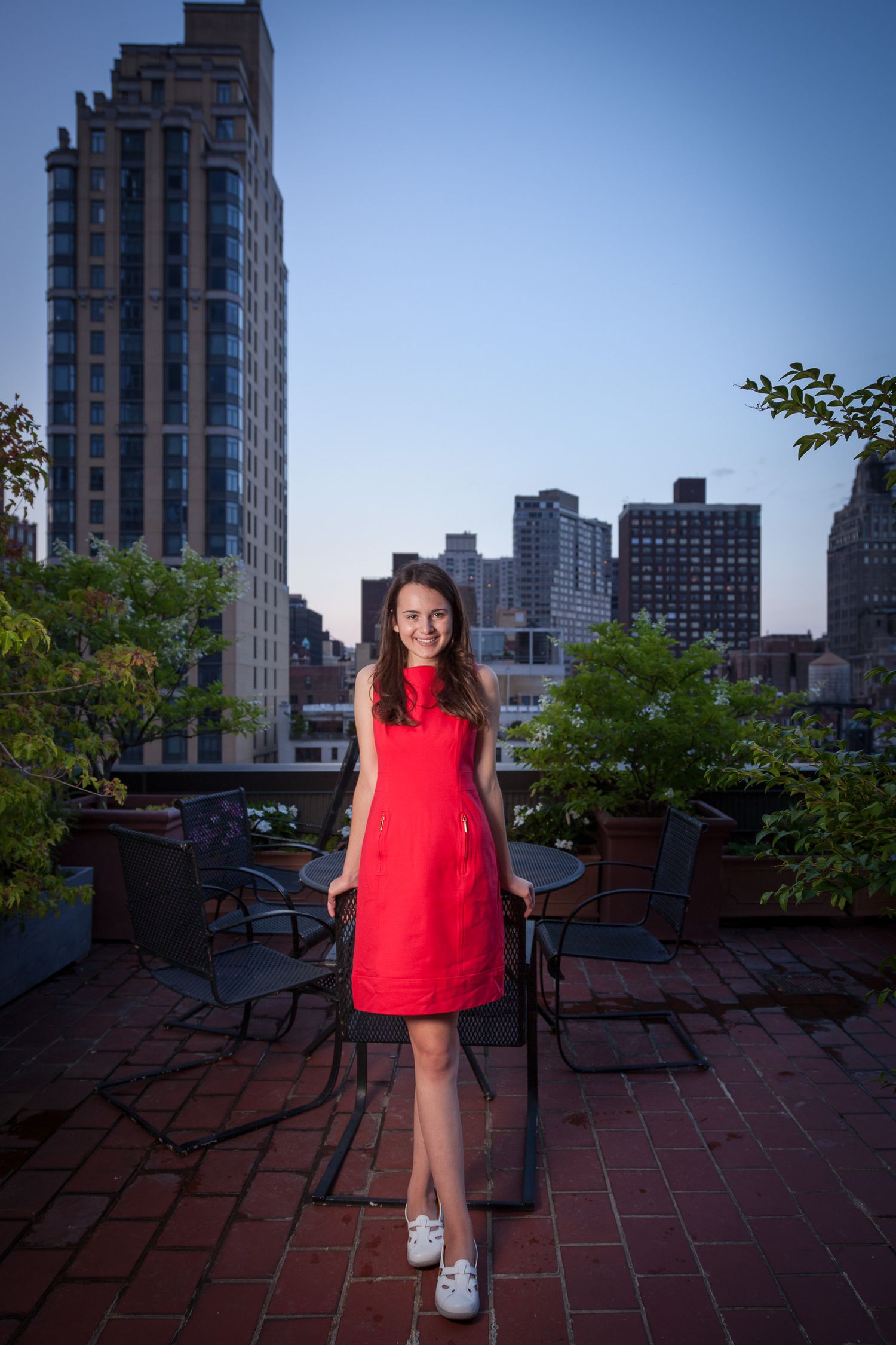 Woman in red dress standing on rooftop terrace with NYC skyline at dusk.