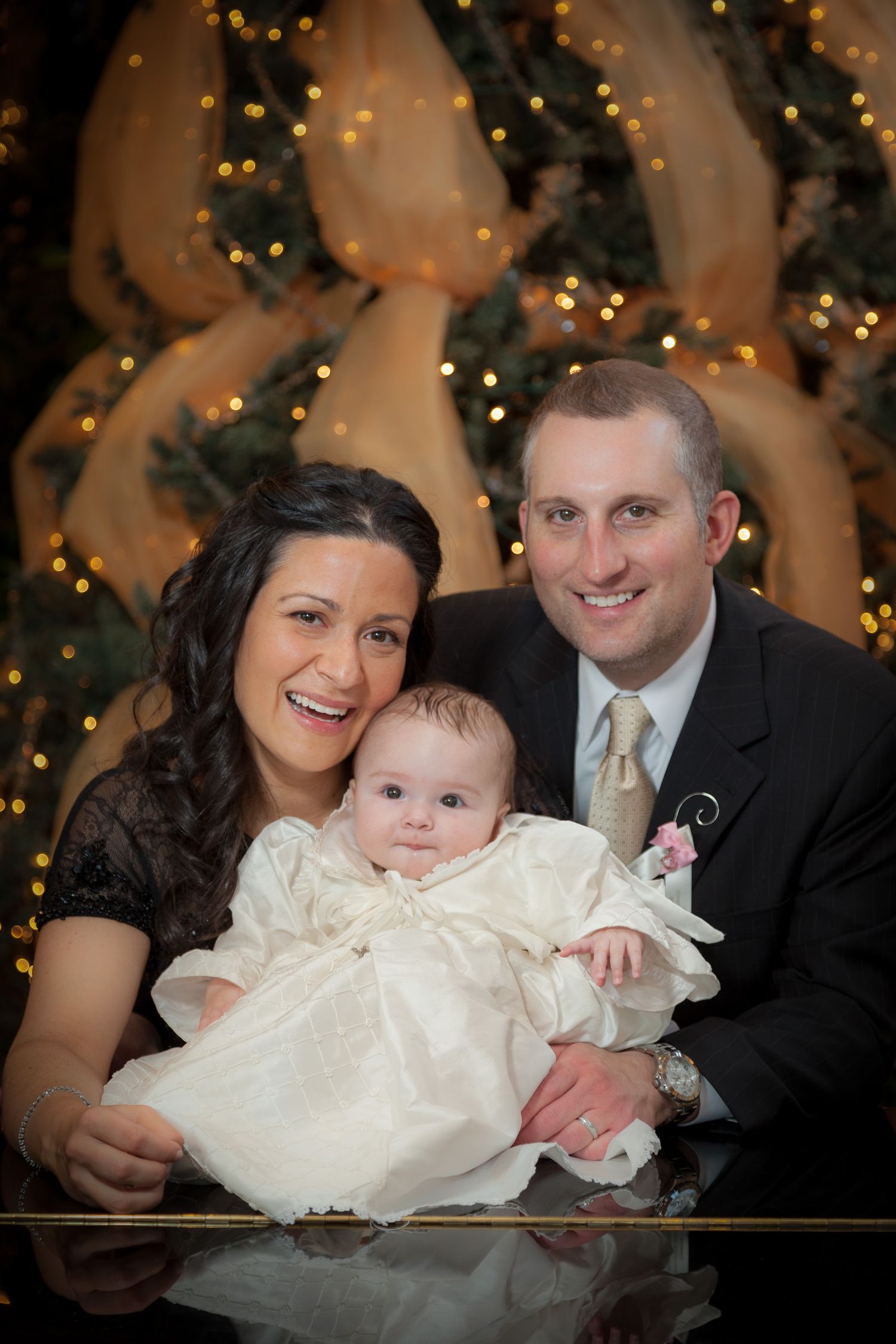 Family portrait of smiling parents with baby in white christening gown near Christmas tree.
