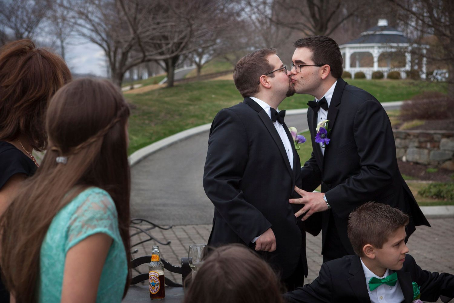 Two grooms share a kiss at their wedding ceremony outdoors, with guests in formal attire surrounding them.