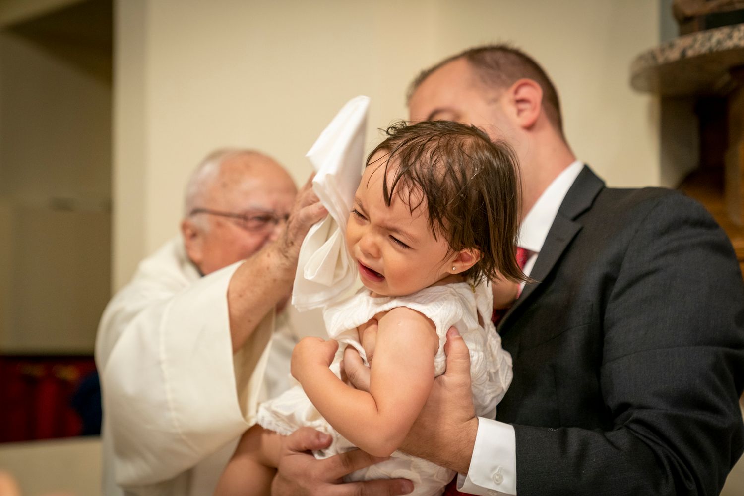 A baby crying during a baptism ceremony held by a priest while a man in a suit holds the infant.