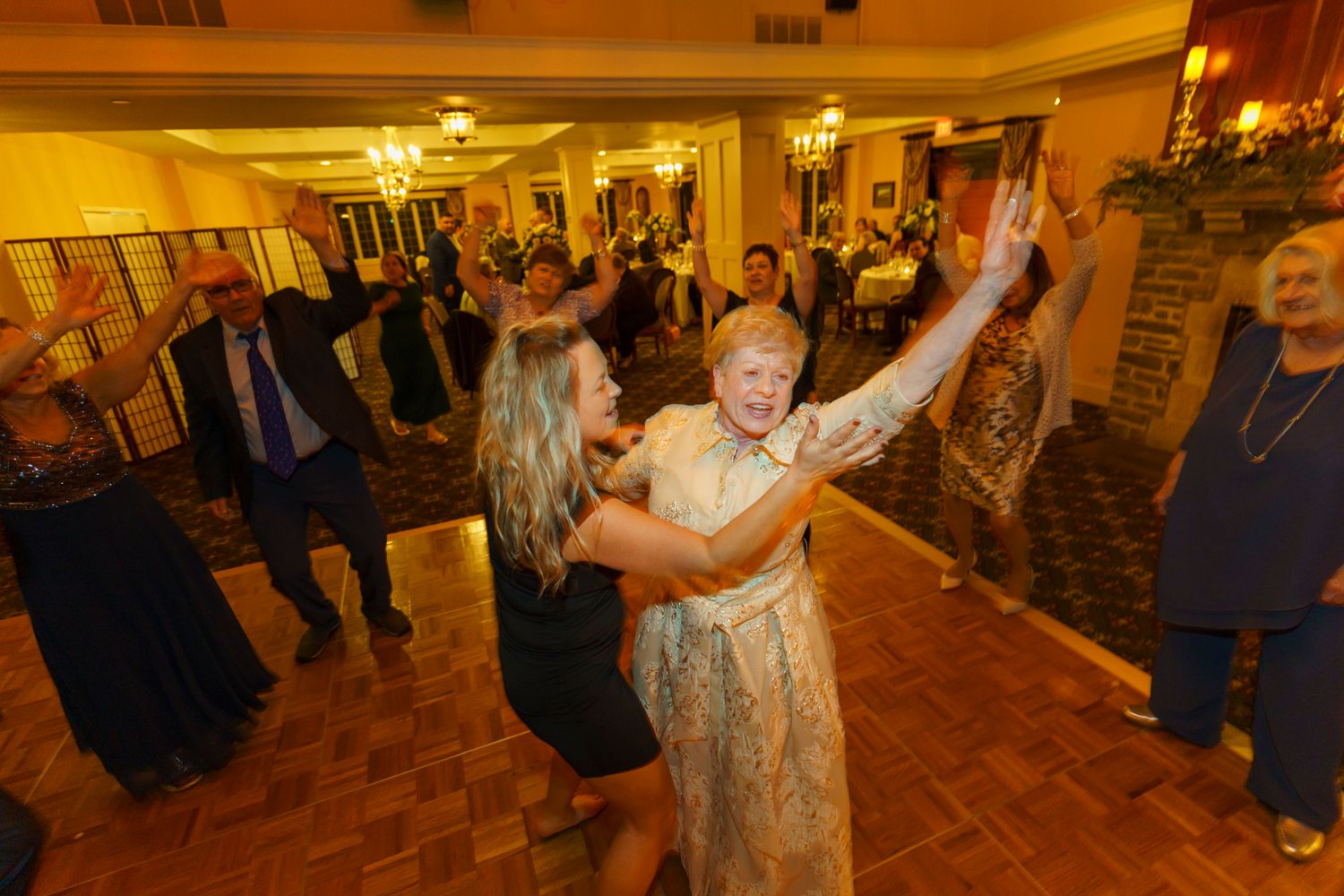 Two women dancing joyfully at an elegant indoor event with guests celebrating in the background.