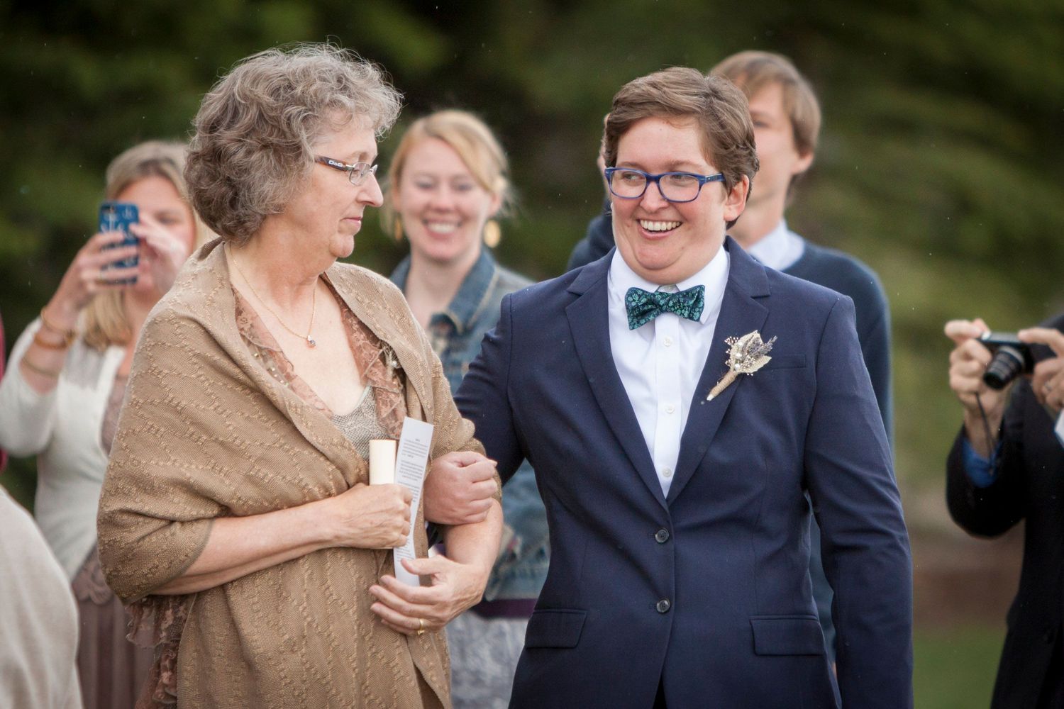 Two people share a joyful moment at an outdoor wedding, one wearing a navy suit with bow tie and boutonniere.