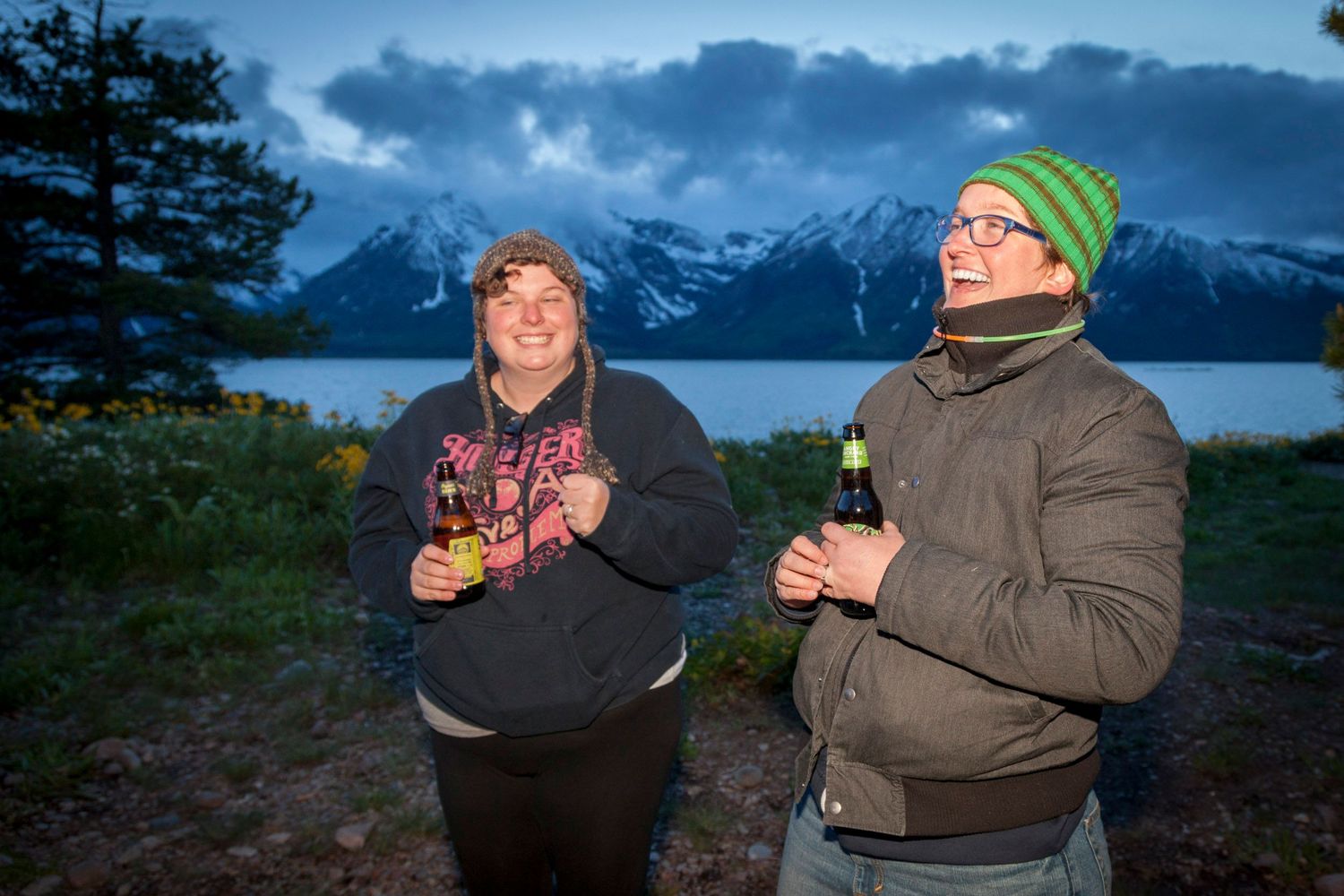Two people laughing outdoors at dusk near a mountain lake with snow-capped peaks in the background.