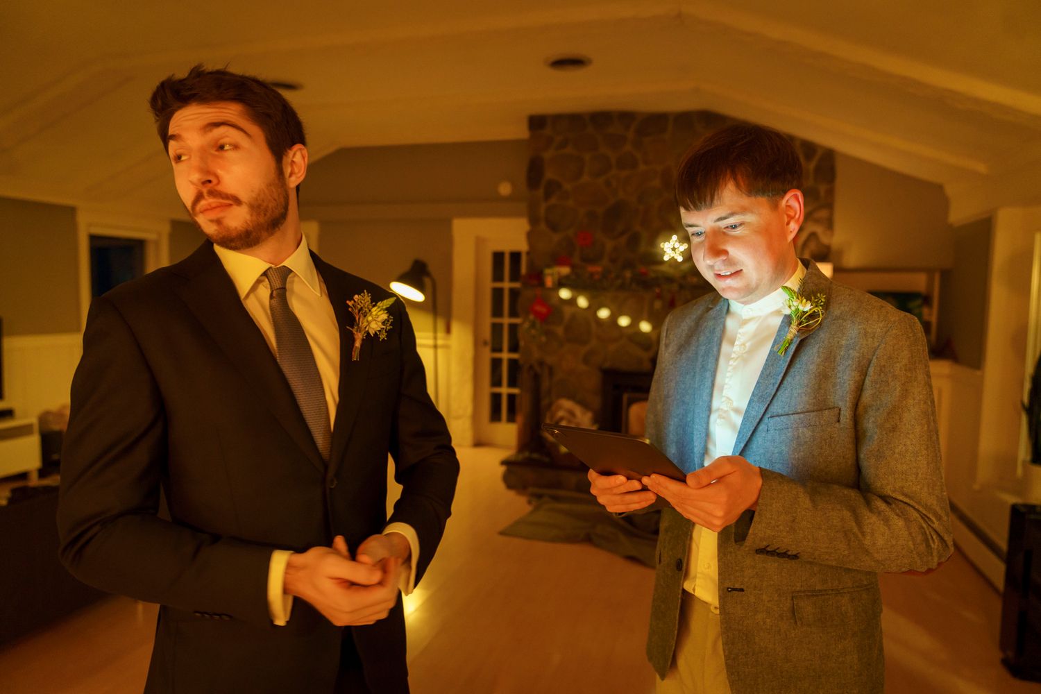 Two men in suits at a wedding, one with a boutonniere looking away, the other checking his phone indoors.