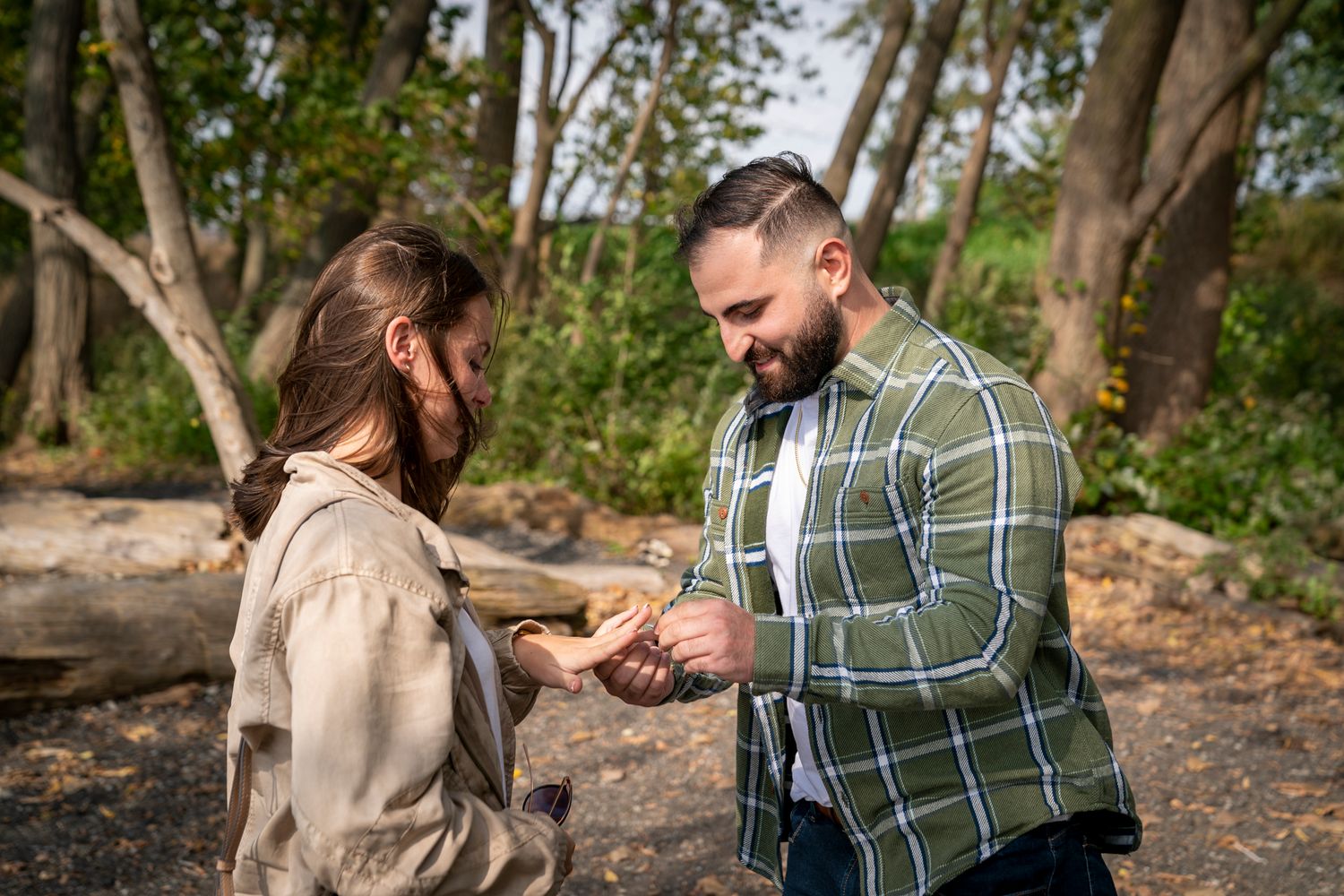 A bearded man in a plaid shirt shares an item with a woman in a forest setting on a sunny day.
