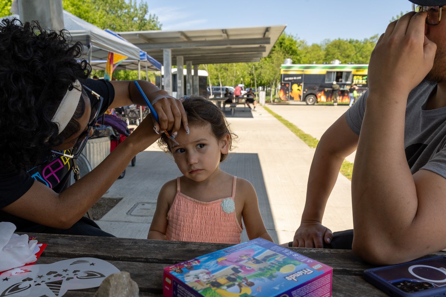 A young girl sits at a picnic table outdoors while an adult braids her hair near a colorful board game.