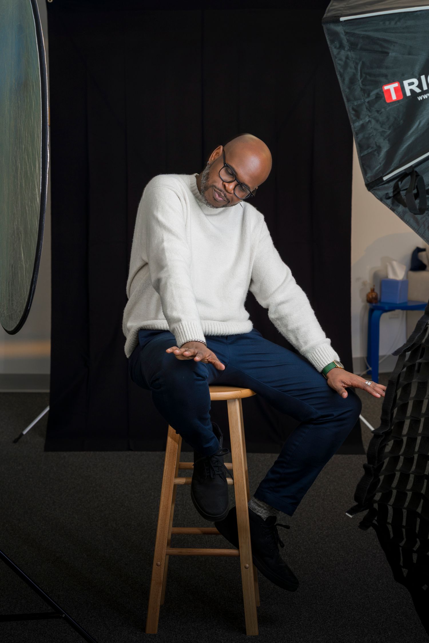 Bald man in white sweater and navy pants sits on wooden stool in professional photo studio with lighting equipment.