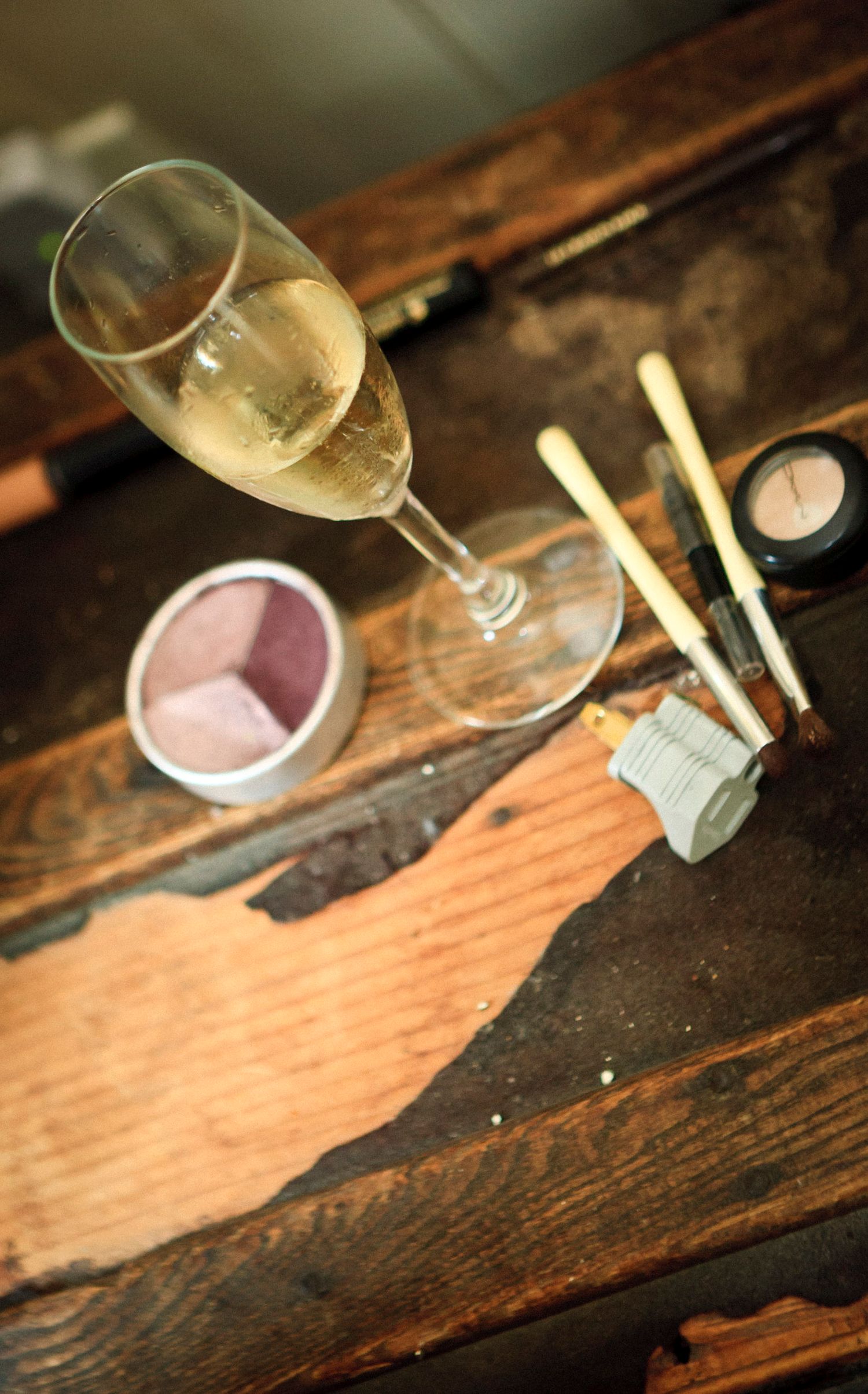 Champagne flute beside makeup brushes and cosmetics on a rustic wooden vanity table.