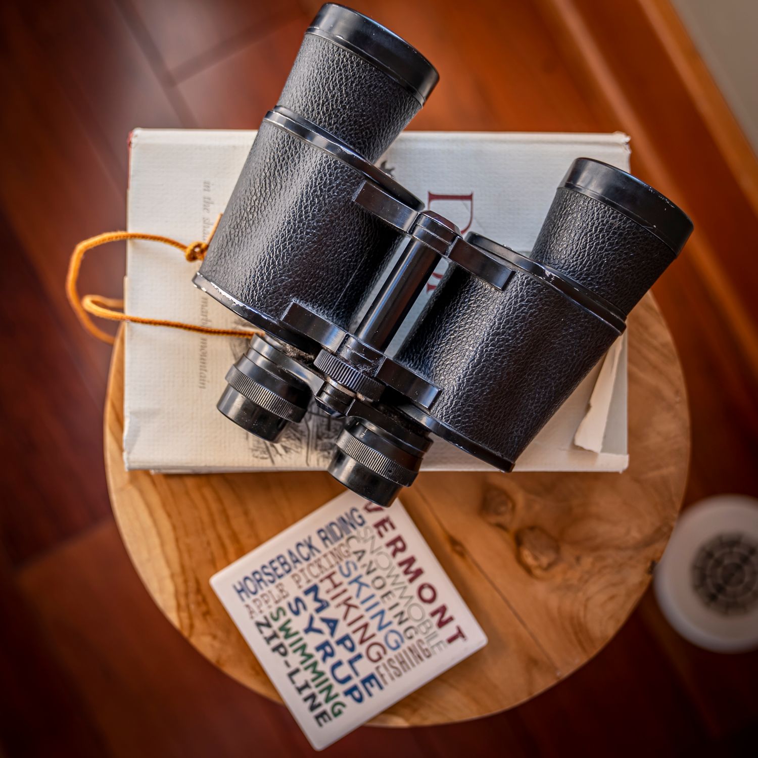 Vintage black binoculars resting on books and a Vermont travel guide on a rustic wooden side table.