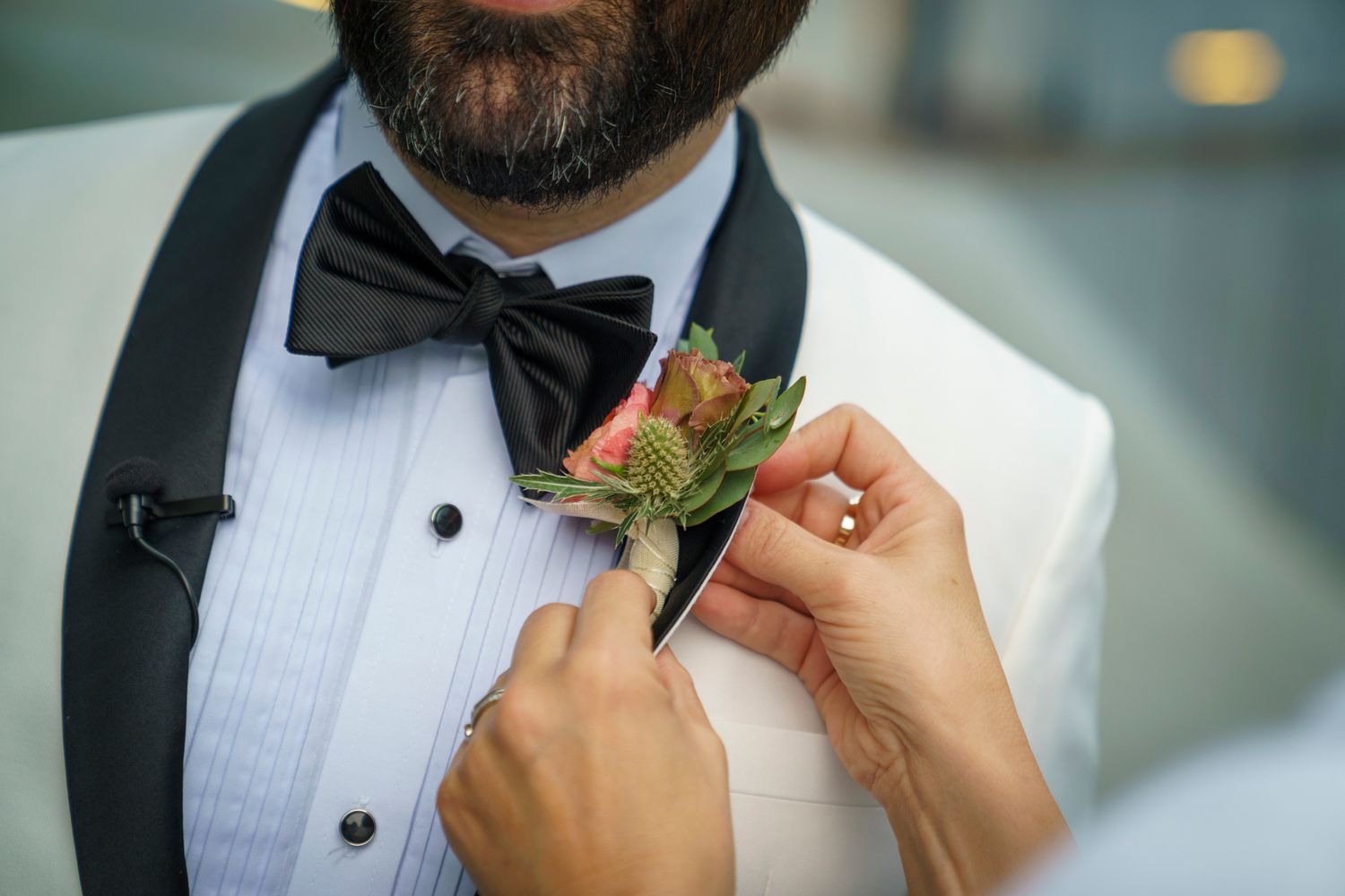 Hands pinning a pink floral boutonniere on a groom wearing a white tuxedo with black bow tie.