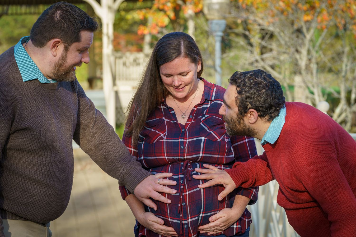 Two men admiring a pregnant woman's baby bump outdoors during a fall maternity photoshoot.