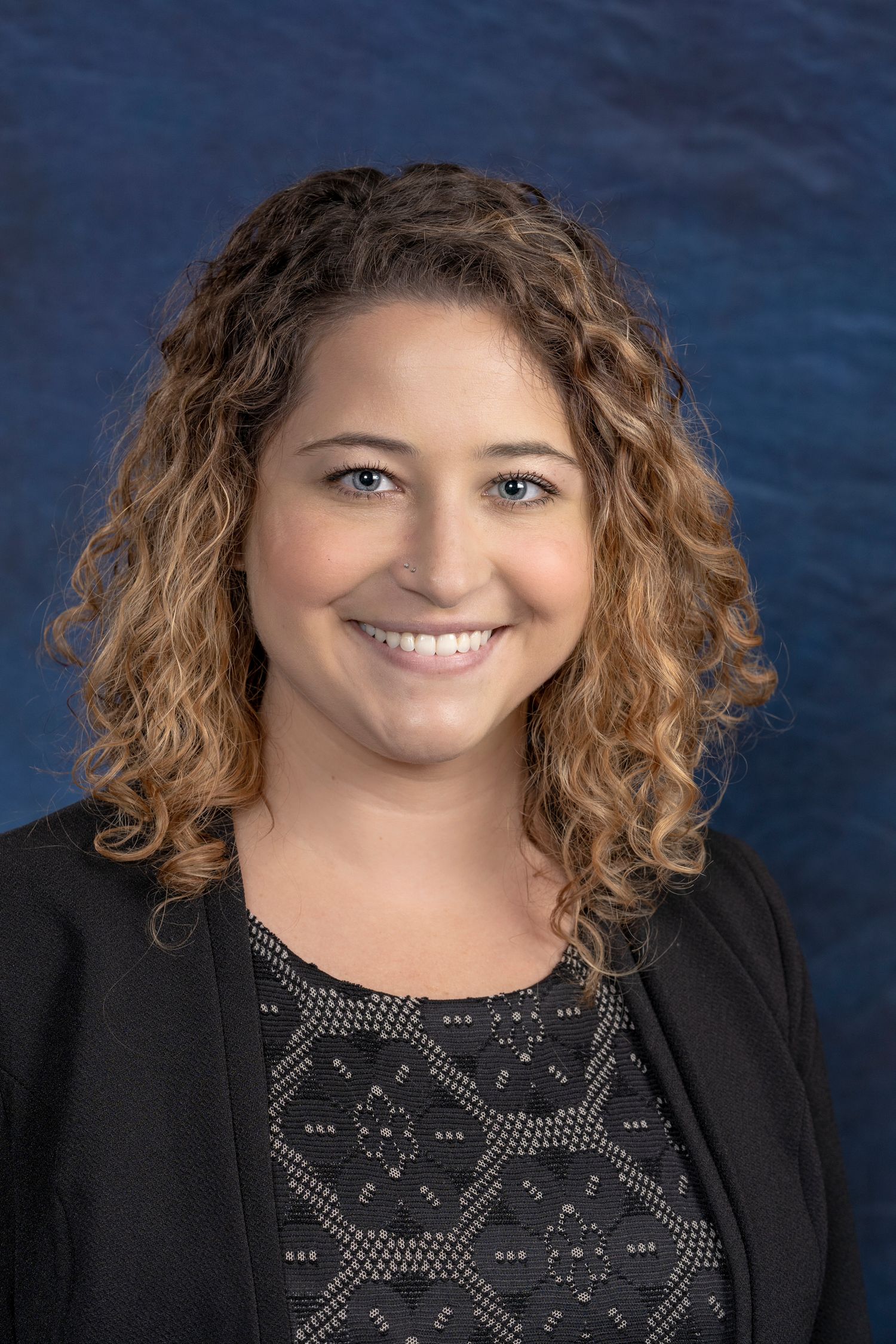 Professional headshot of a smiling woman with curly auburn hair wearing a black blazer against a blue background.