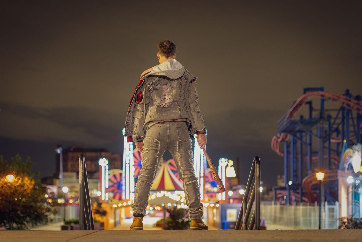 Man in a gray hoodie and jeans stands with his back to the camera, facing a brightly lit amusement park and roller coaster at night.