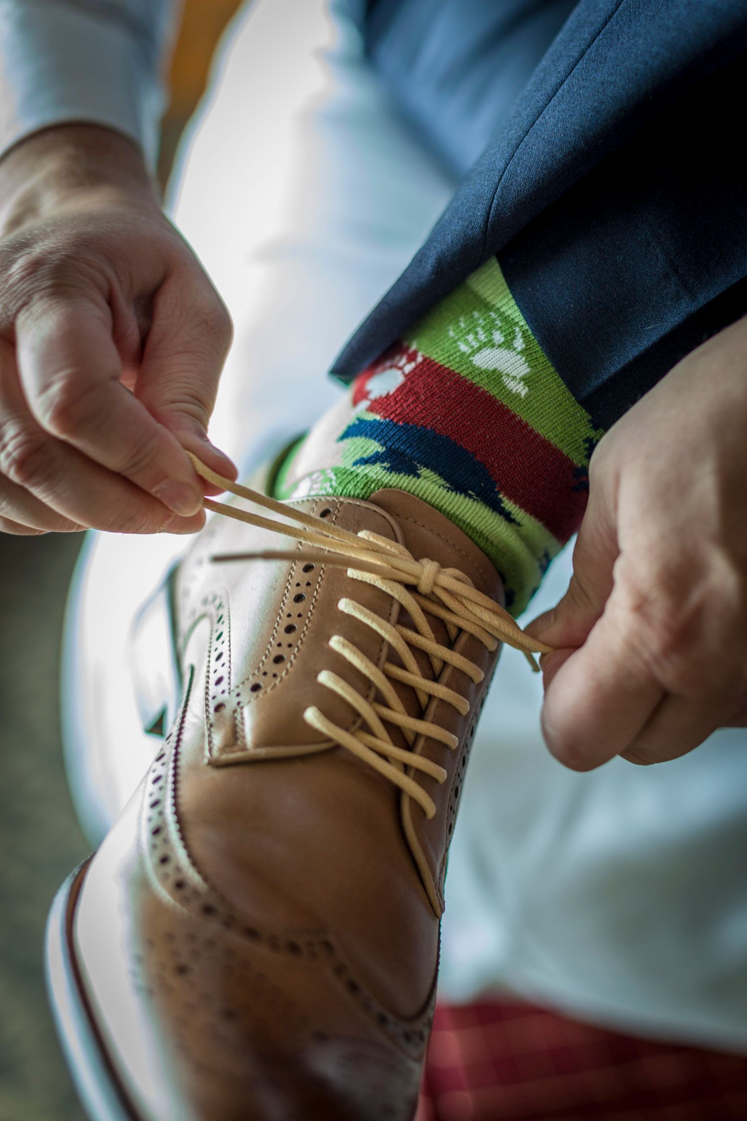Close-up of groom tying tan brogue dress shoes with colorful holiday socks during wedding preparations.