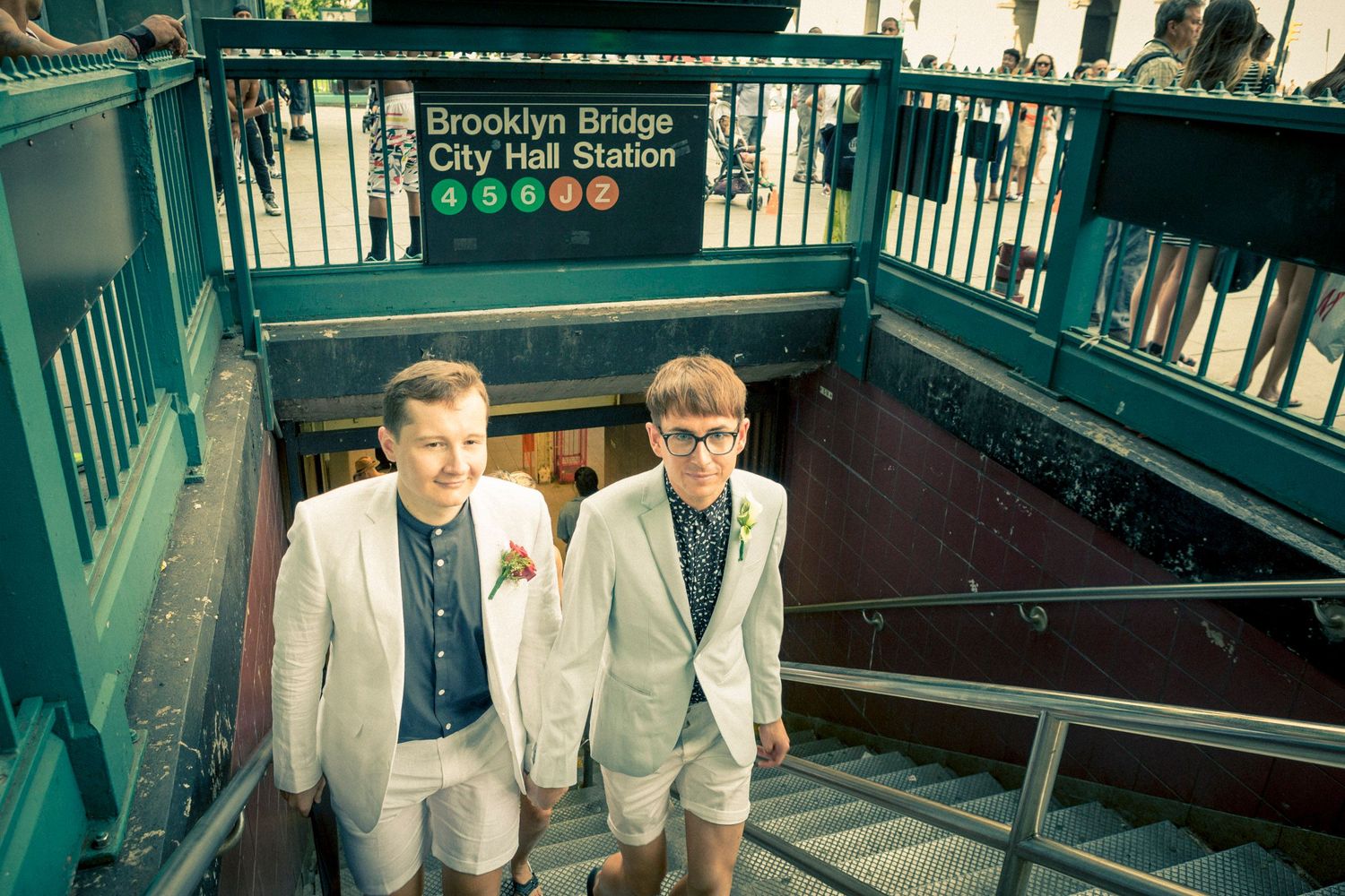 Two men in white suits pose on subway stairs at Brooklyn Bridge City Hall Station in New York City.