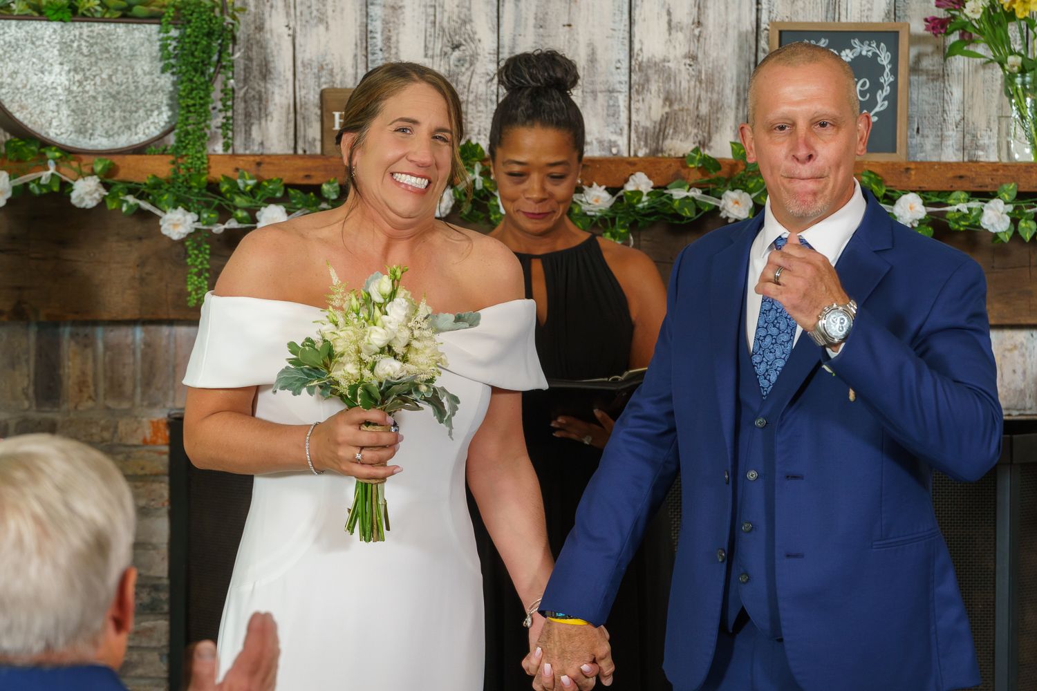 Happy couple holding hands at wedding ceremony with officiant, bride in white gown holding bouquet, groom in blue suit.