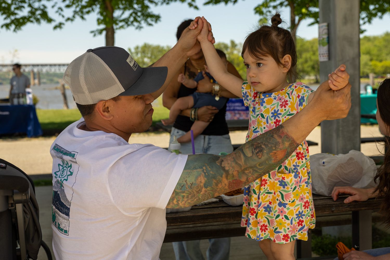 Tattooed man in gray cap gives high five to young girl in floral dress at outdoor park event.