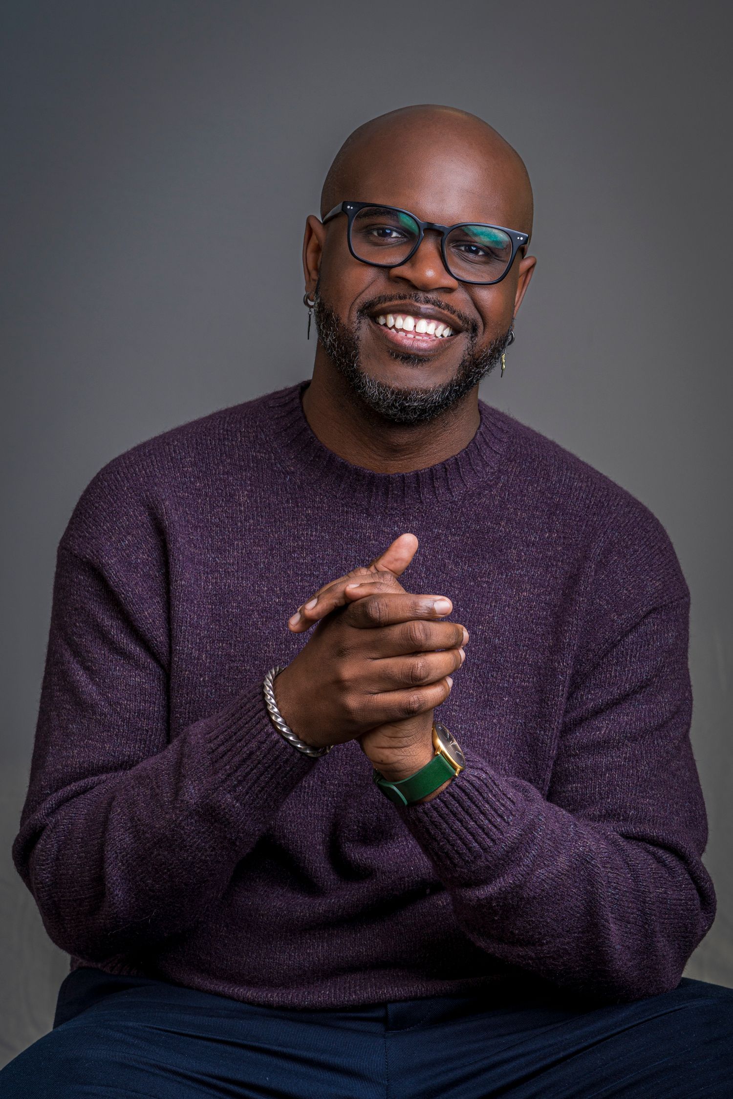Smiling bald Black man with glasses and beard wearing purple sweater, hands clasped, studio portrait.