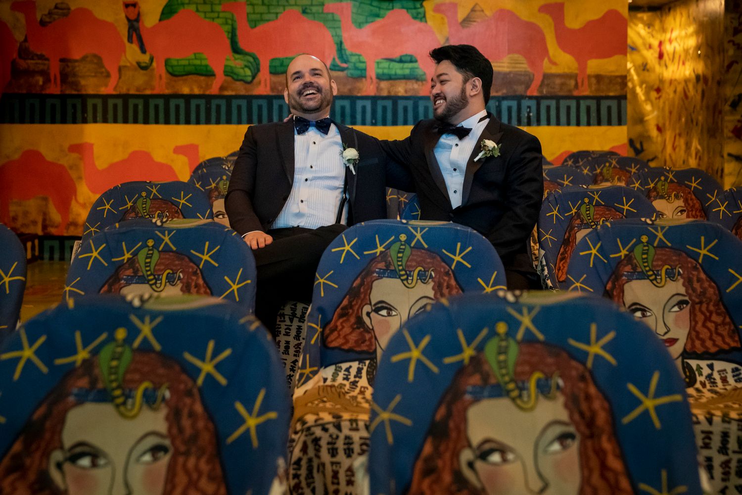 Two grooms in tuxedos share a joyful moment seated among ornate painted chairs at their wedding reception.