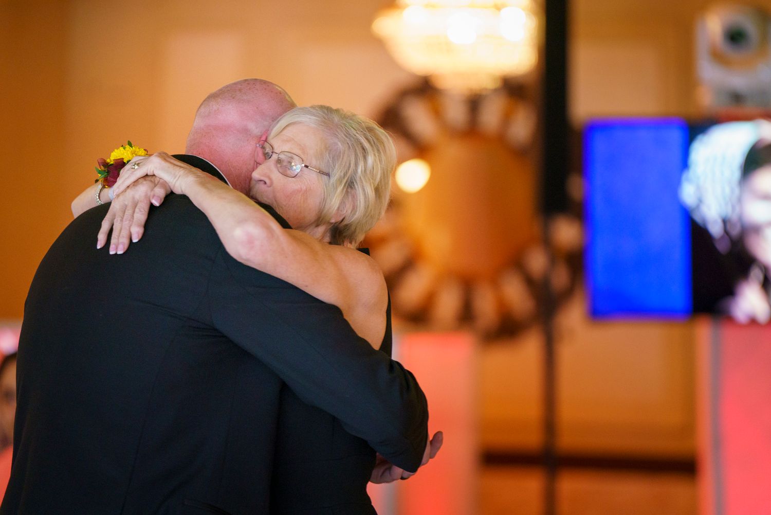 Emotional wedding reception embrace between two guests near a sunflower centerpiece and DJ setup.