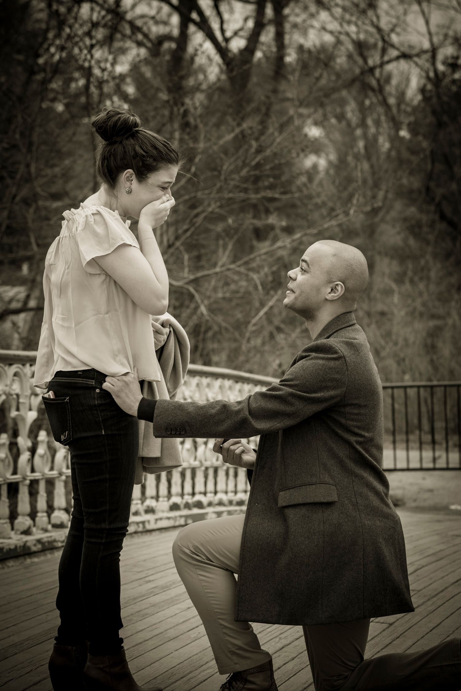 A man kneels on one knee proposing to a tearful woman on a wooden deck in a romantic black and white outdoor scene.