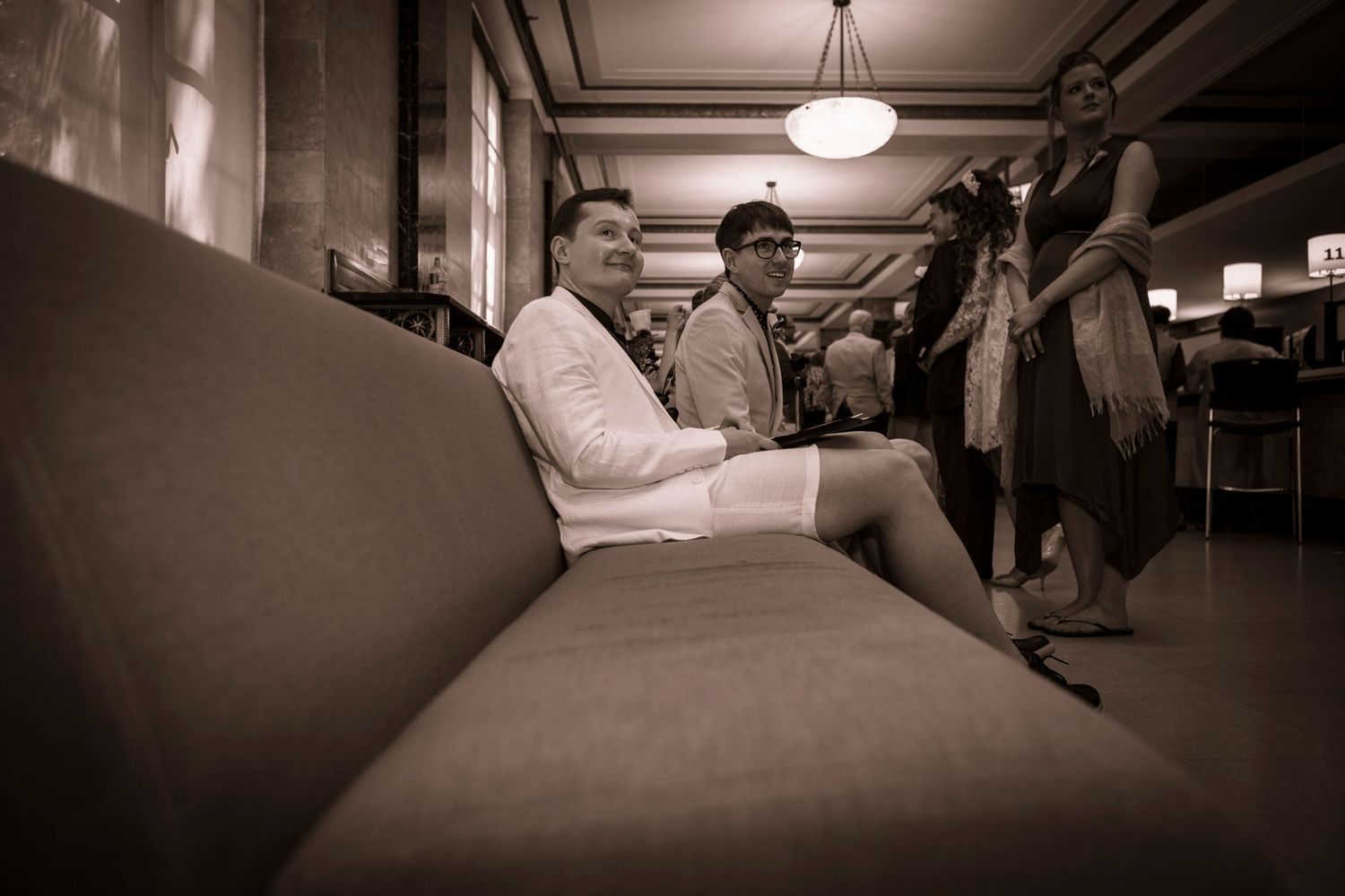 Two people sitting on a couch in a vintage-style lobby, captured in sepia tone photography.