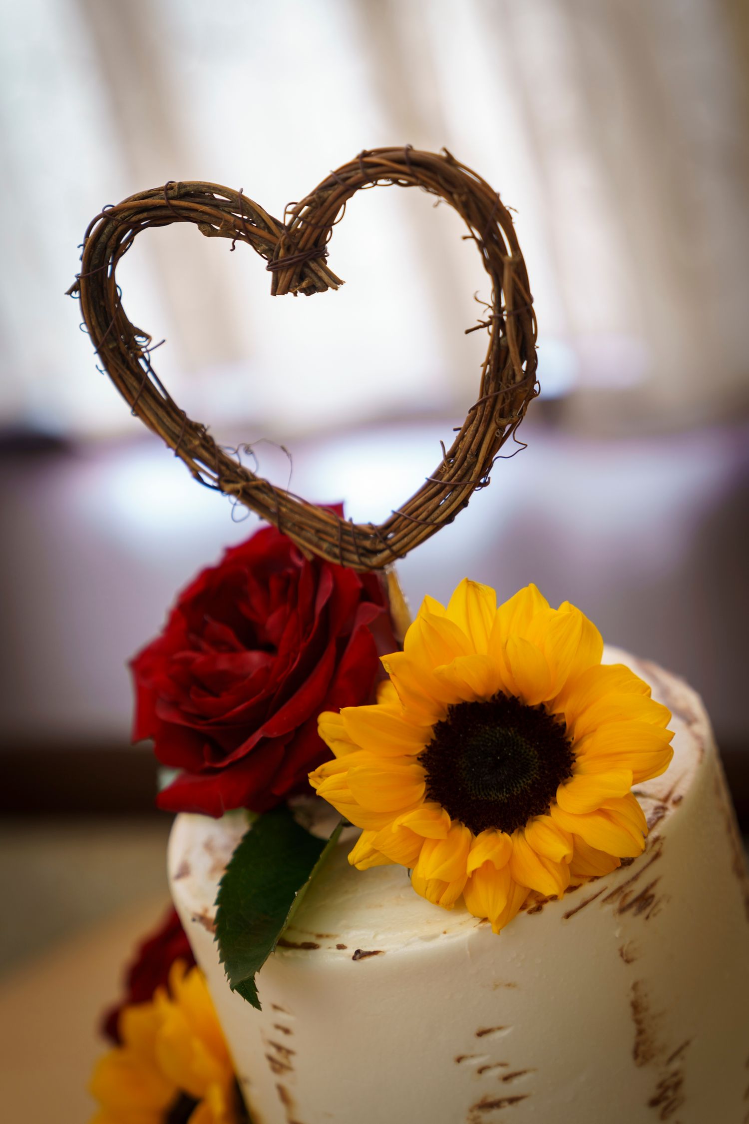 Rustic wedding cake with twig heart topper, red rose, and sunflower decoration on white frosted tiered cake.