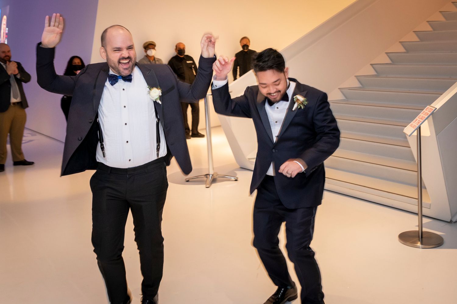 Two grooms celebrate joyfully at their wedding reception, raising arms and dancing in elegant tuxedos.