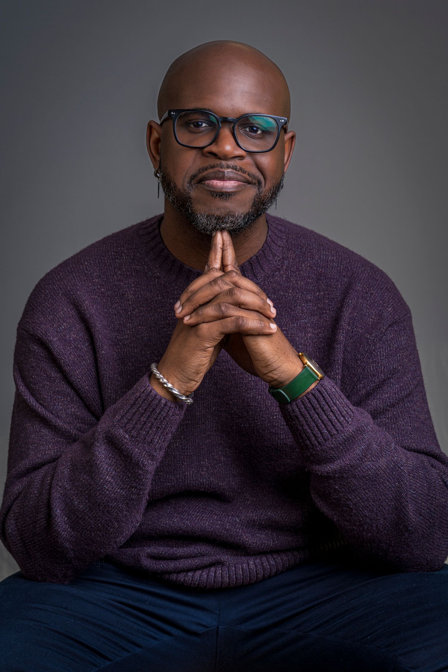 Confident bald Black man with glasses and beard, wearing purple sweater, hands clasped, studio portrait.