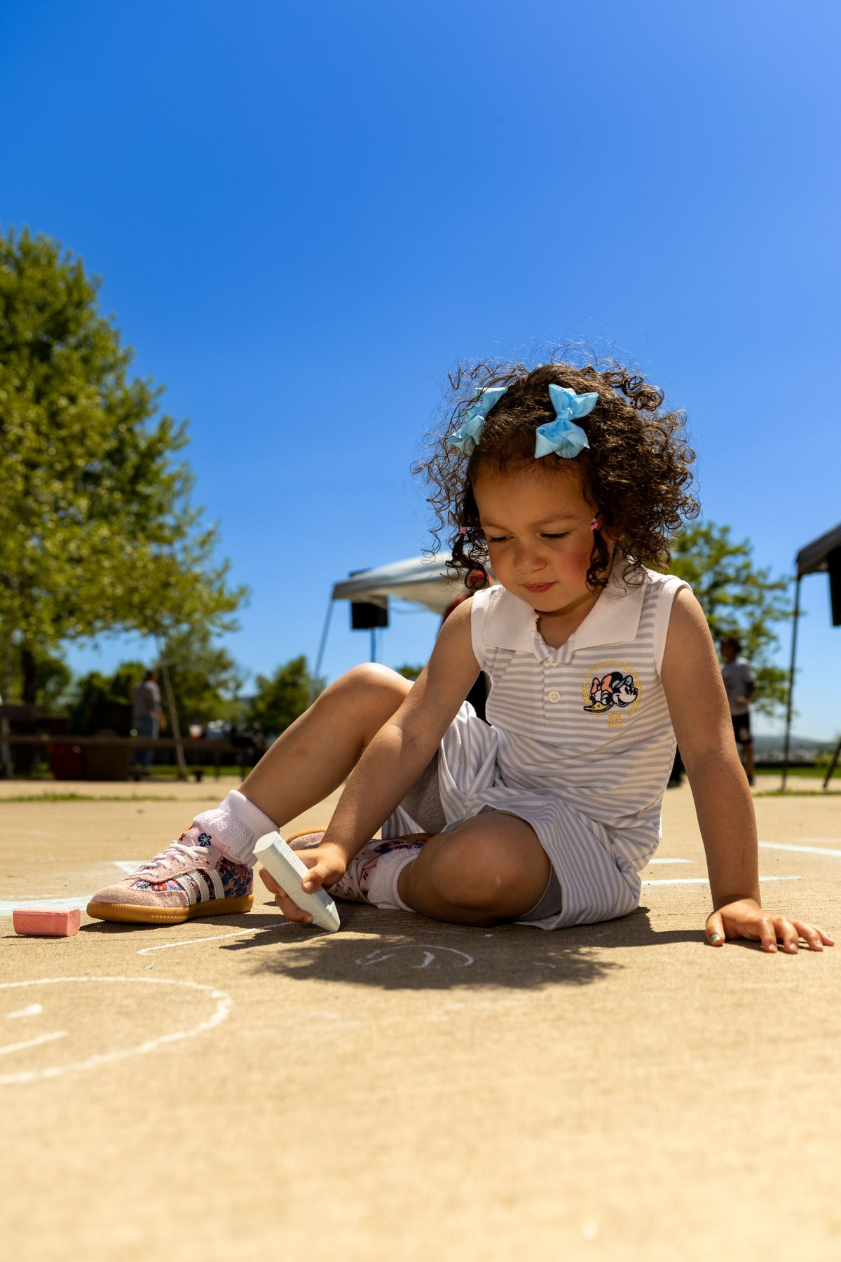 Young curly-haired girl sitting on pavement drawing with chalk on a sunny day outdoors.