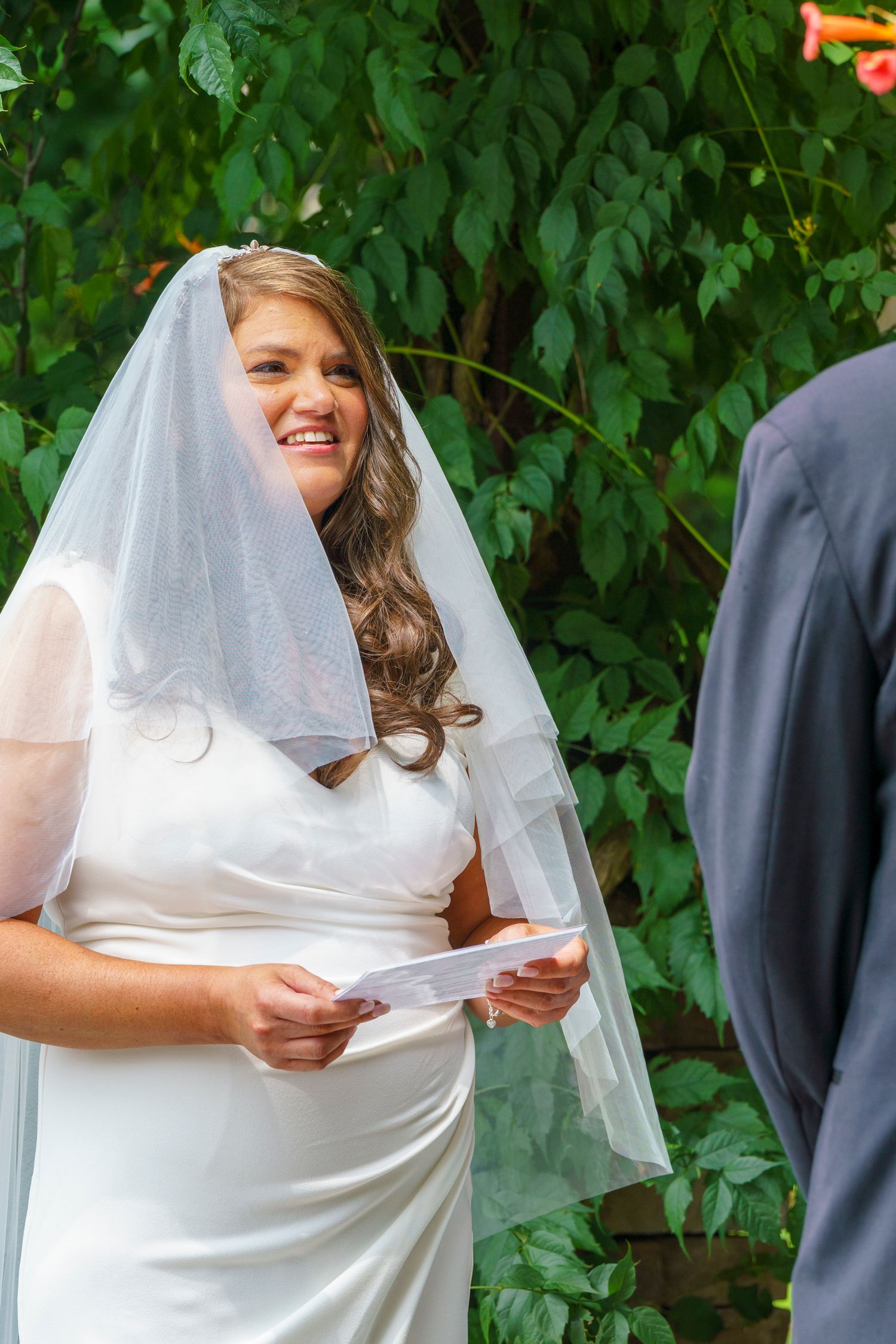 Smiling bride in white gown and veil reads wedding vows outdoors, with lush green foliage in the background.