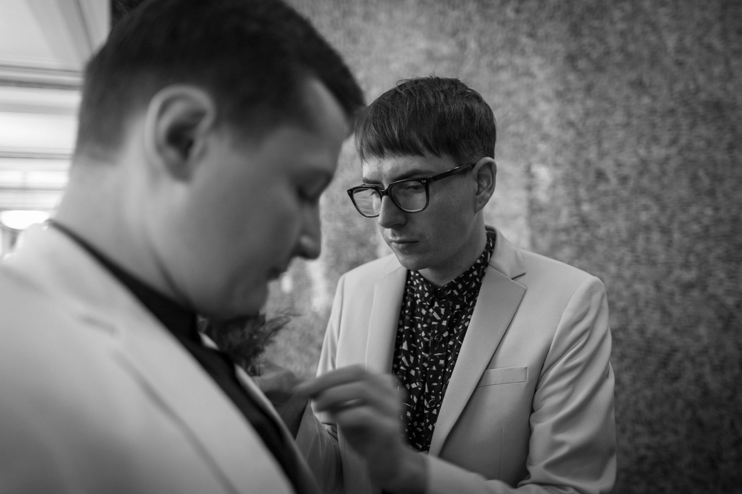 Black and white photo of two men in suits, one adjusting the other's boutonniere at a formal event.