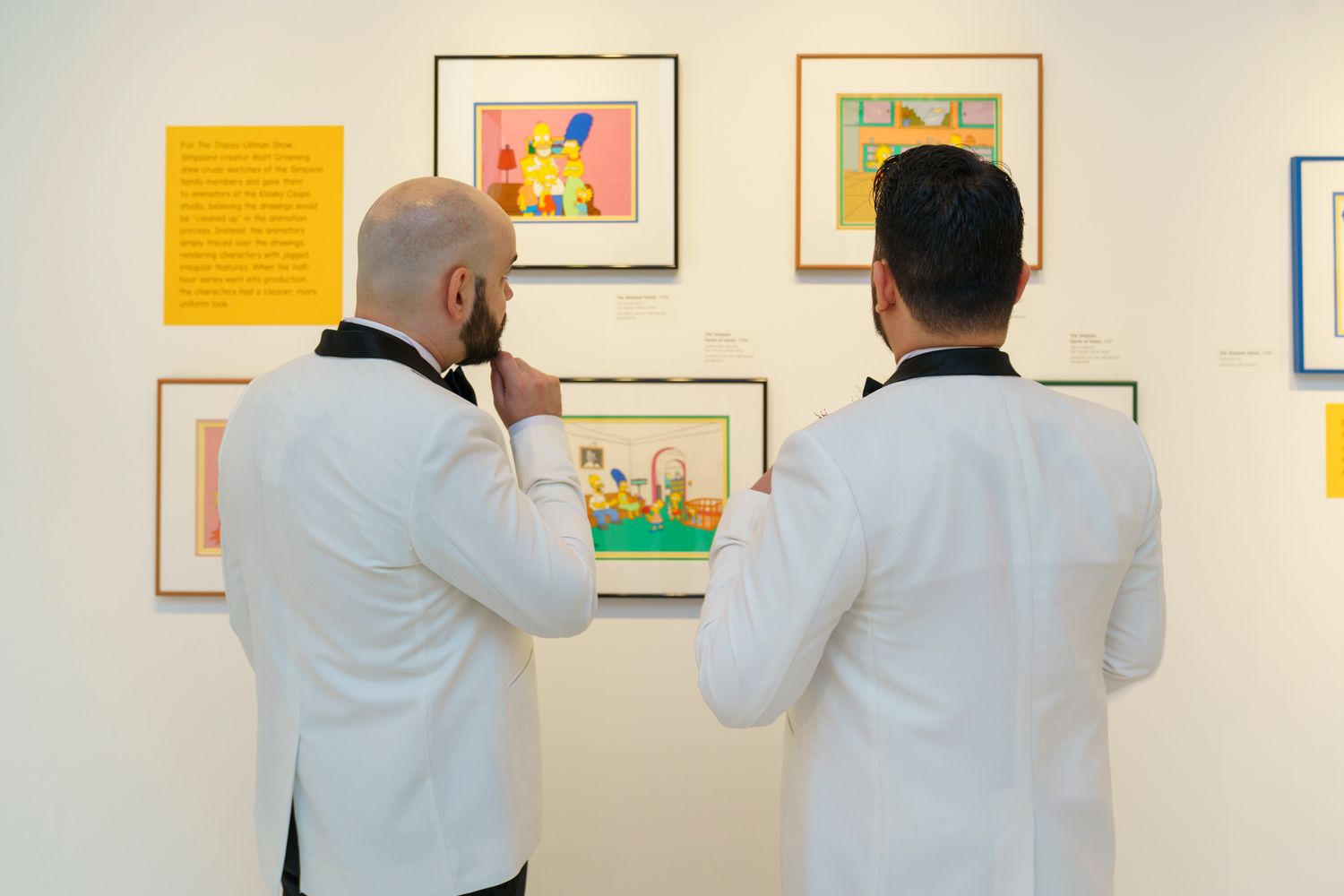Two grooms in white tuxedos viewing Simpsons artwork at gallery during their wedding celebration.