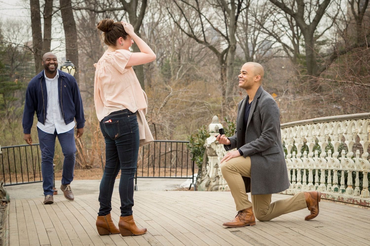 Man kneeling on one knee proposing to a surprised woman on a wooden deck in a park setting.