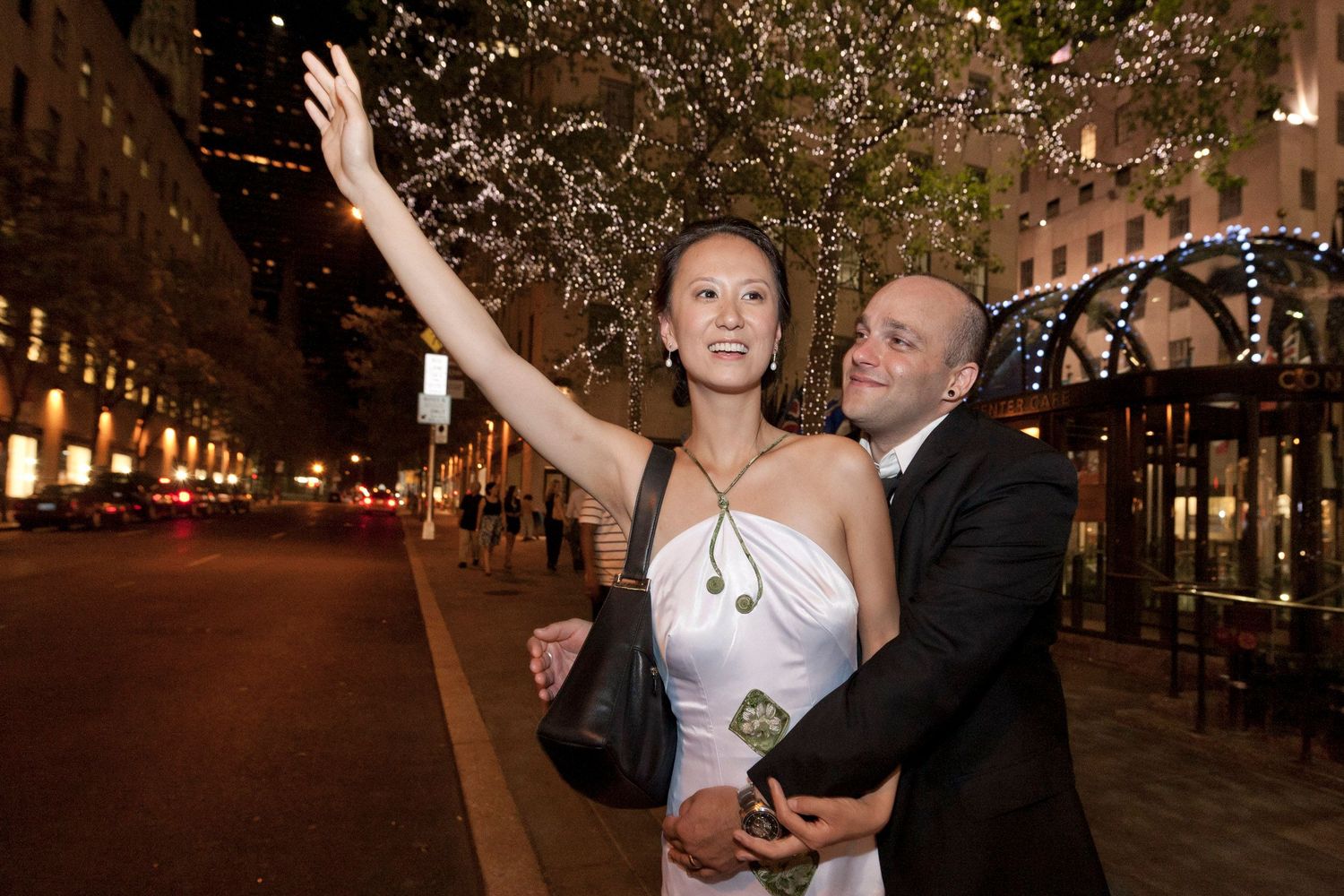 Newlywed couple in wedding attire hails a taxi on a city street lit with sparkling lights.