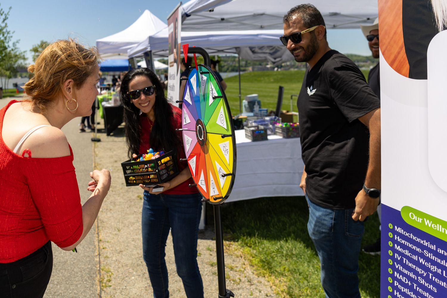 People at outdoor event with colorful prize spin wheel at vendor booth on sunny day.